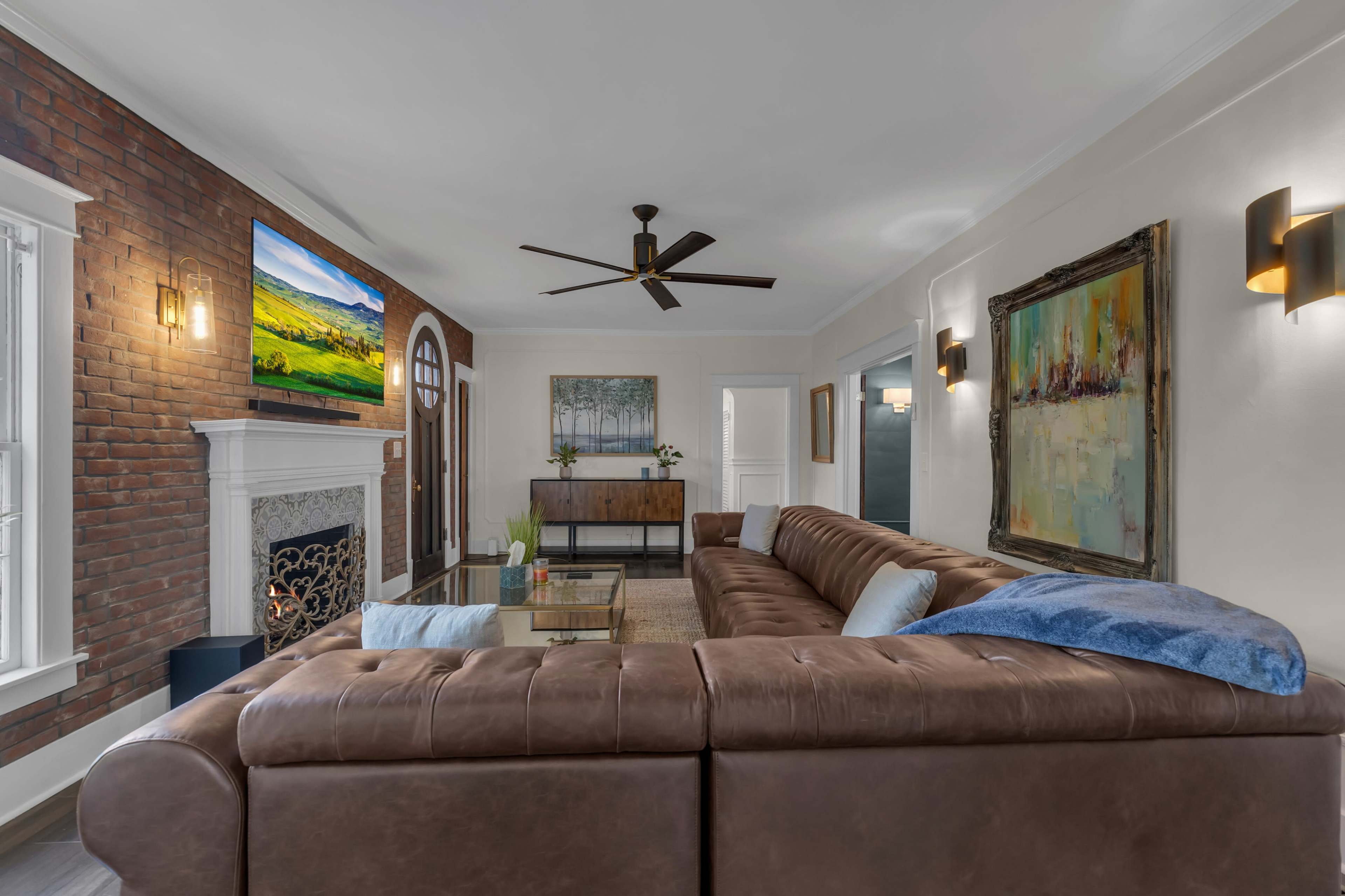 A spacious living room features a large brown leather sectional sofa, a brick accent wall, a mounted television, and artwork above a decorative fireplace.