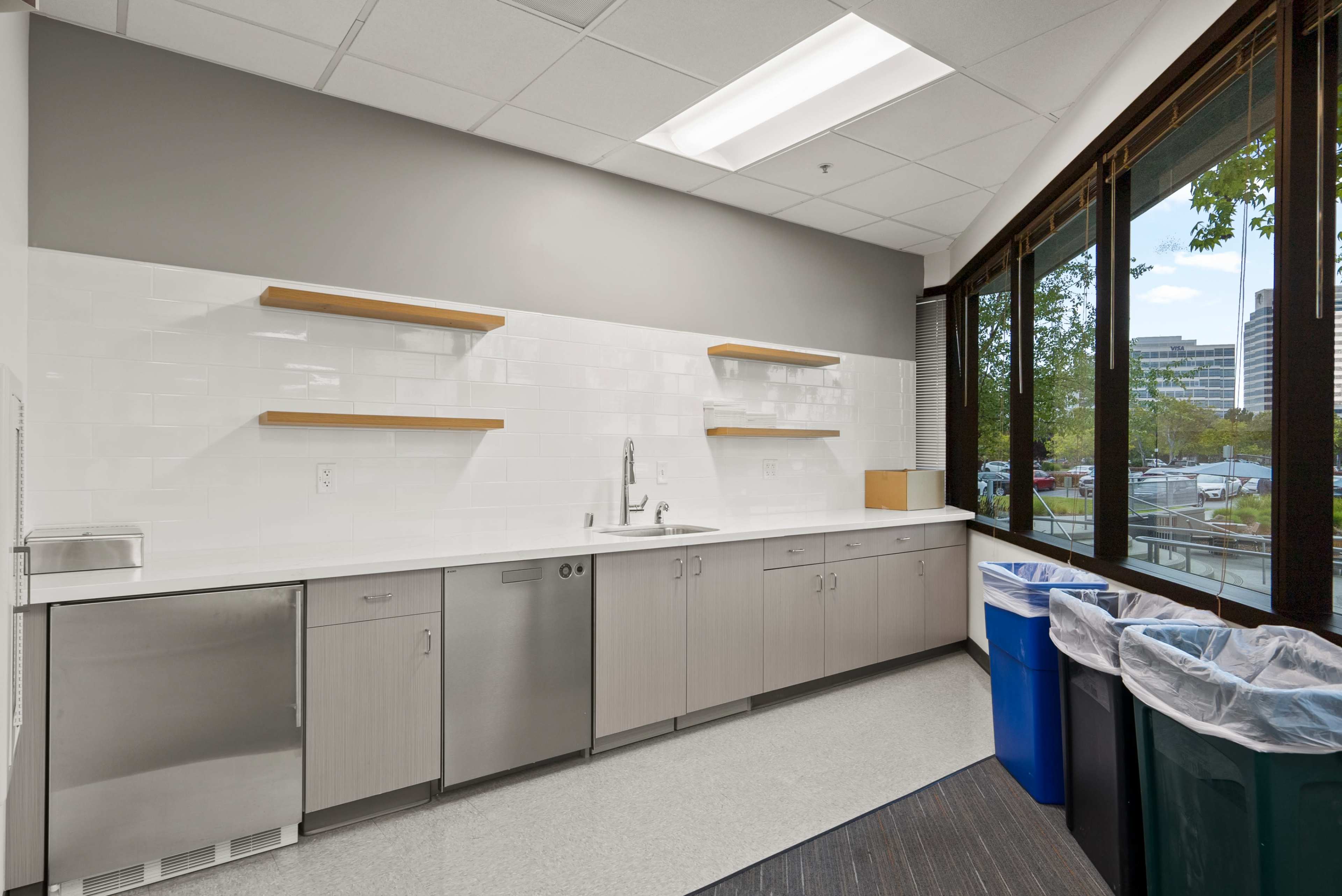 The image shows a clean kitchen area with a sink, cabinets, and open shelves, along with waste bins positioned near a window.