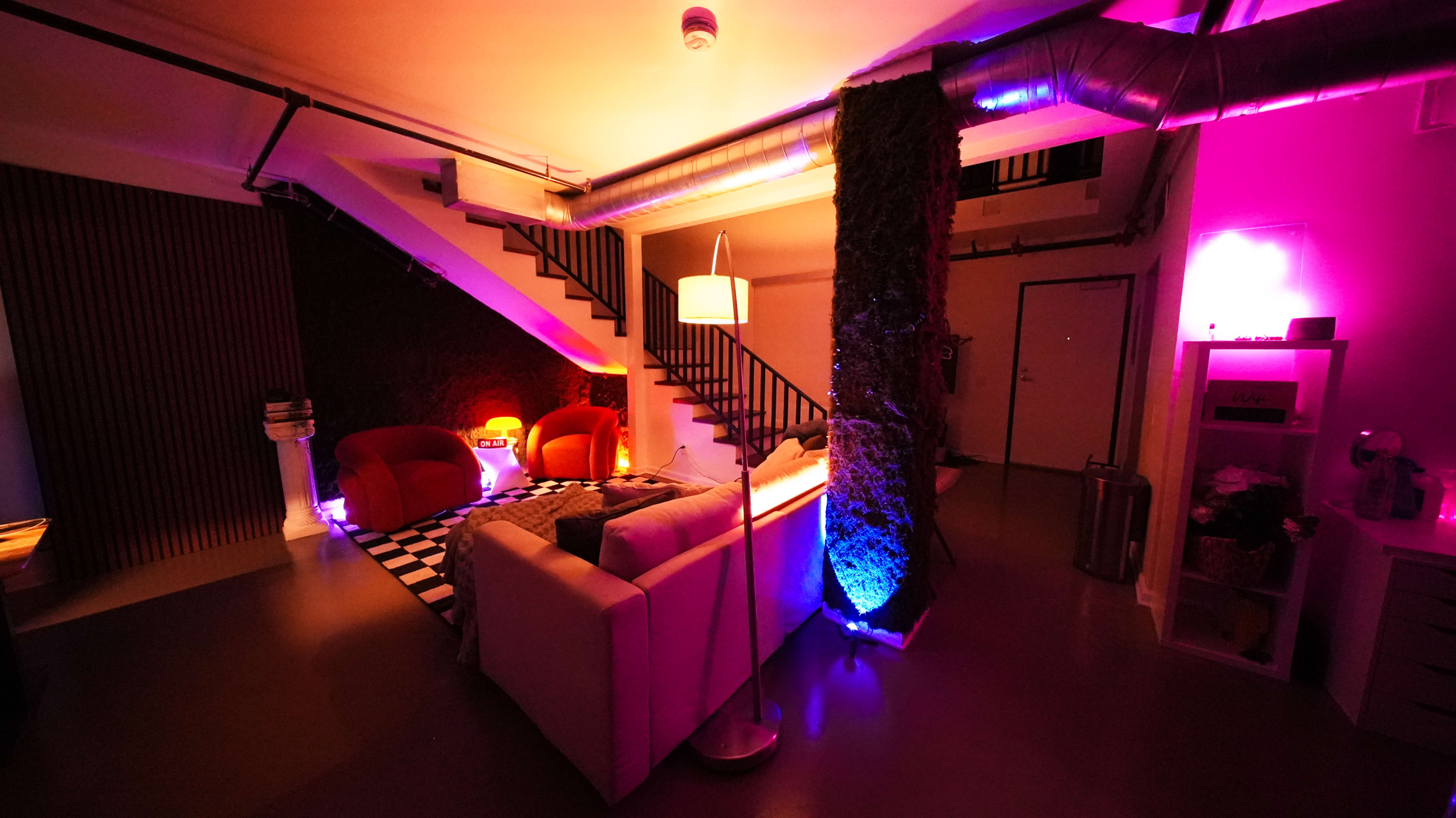The image shows a modern interior space with a staircase, a cozy seating area featuring two red chairs, and colorful lighting along the walls.