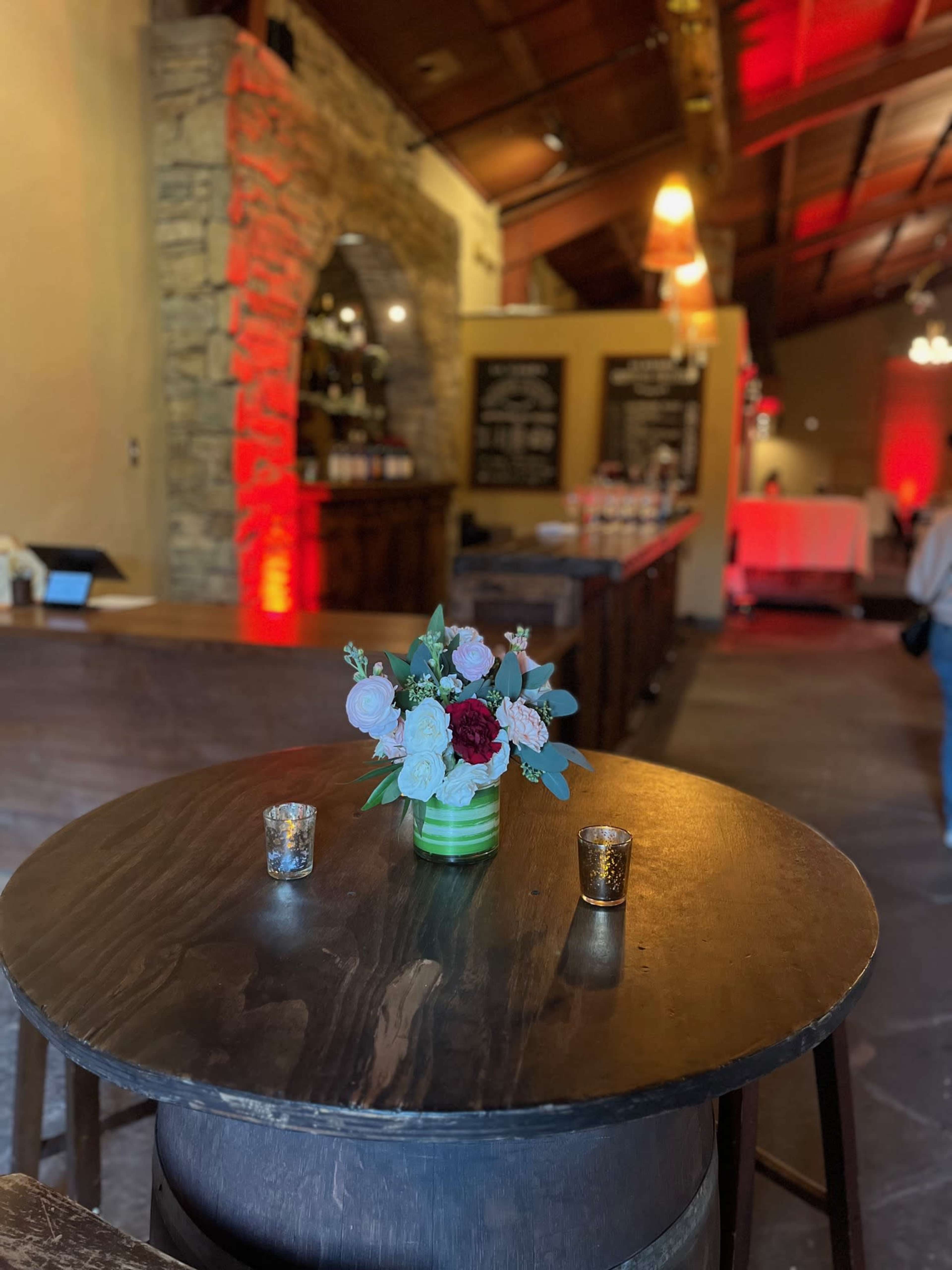 A round wooden table displays a floral arrangement and two candles in a rustic restaurant setting.
