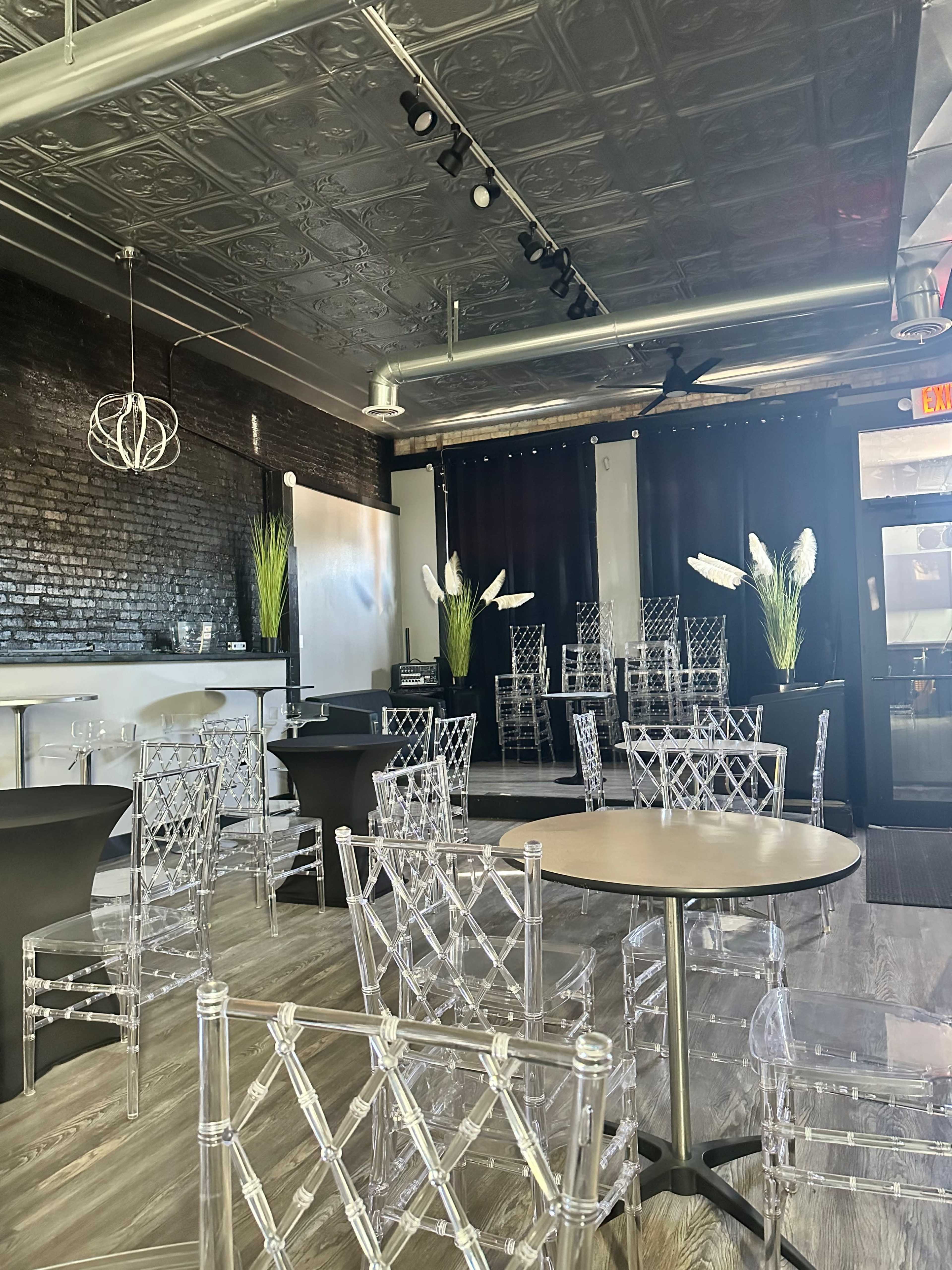 The interior features a modern event space with transparent chairs arranged around tables, dark walls, and decorative plants.