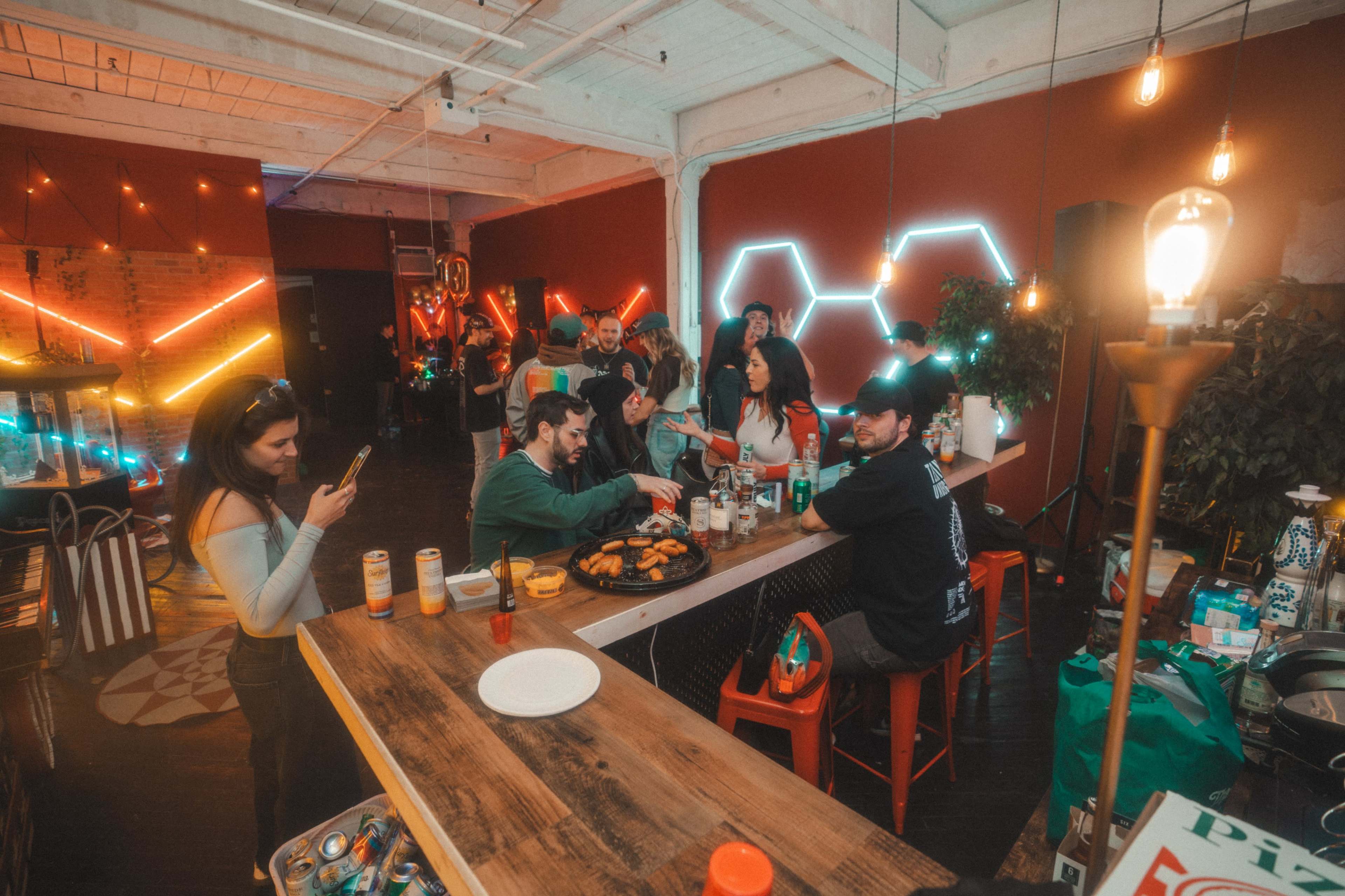 A vibrant gathering occurs in a spacious venue with colorful lighting, where people socialize around a bar and table filled with food and drinks.