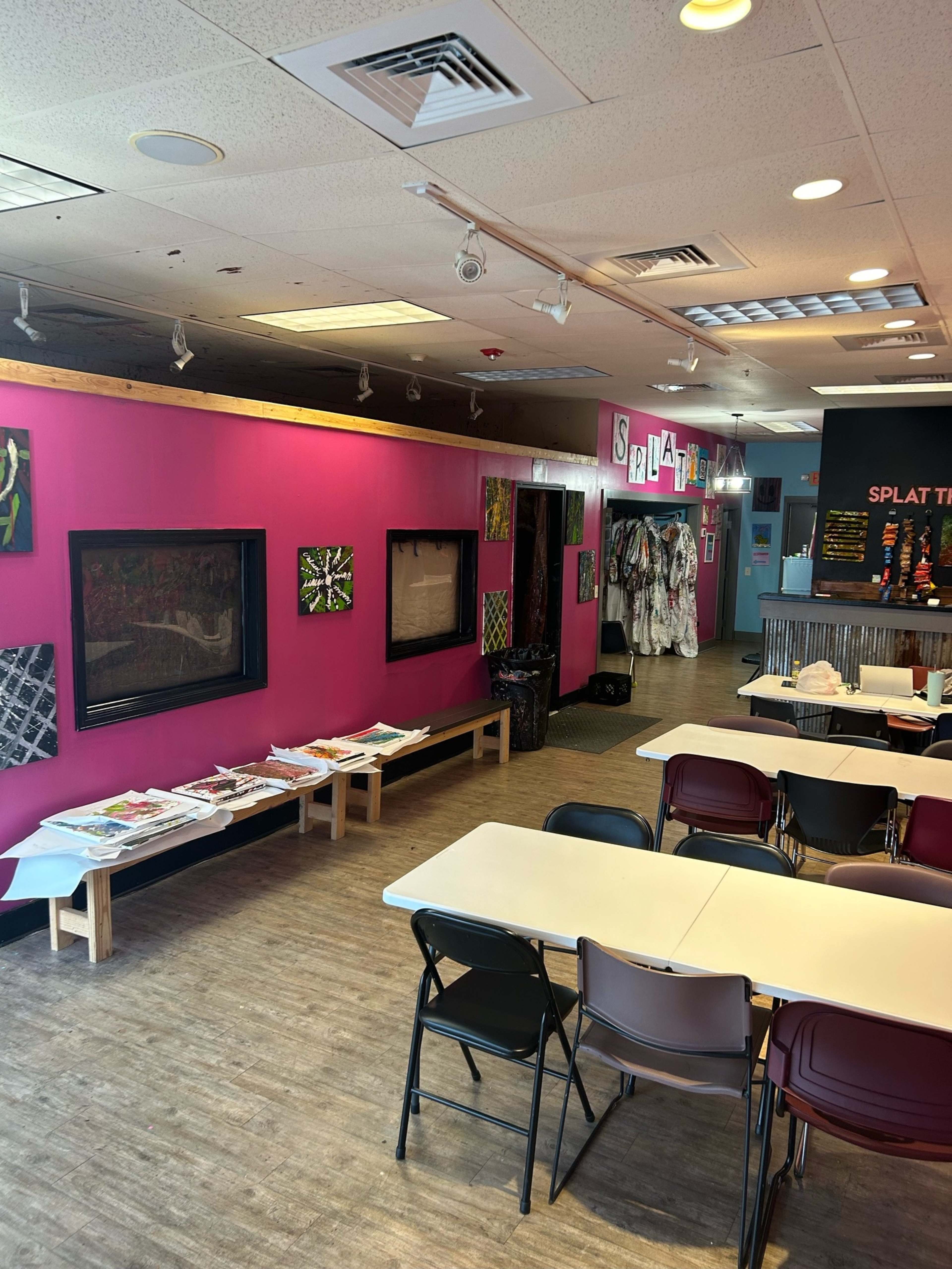 An art studio with bright pink walls, displaying artwork on the walls, tables set up for patrons, and separate areas for creative activities.