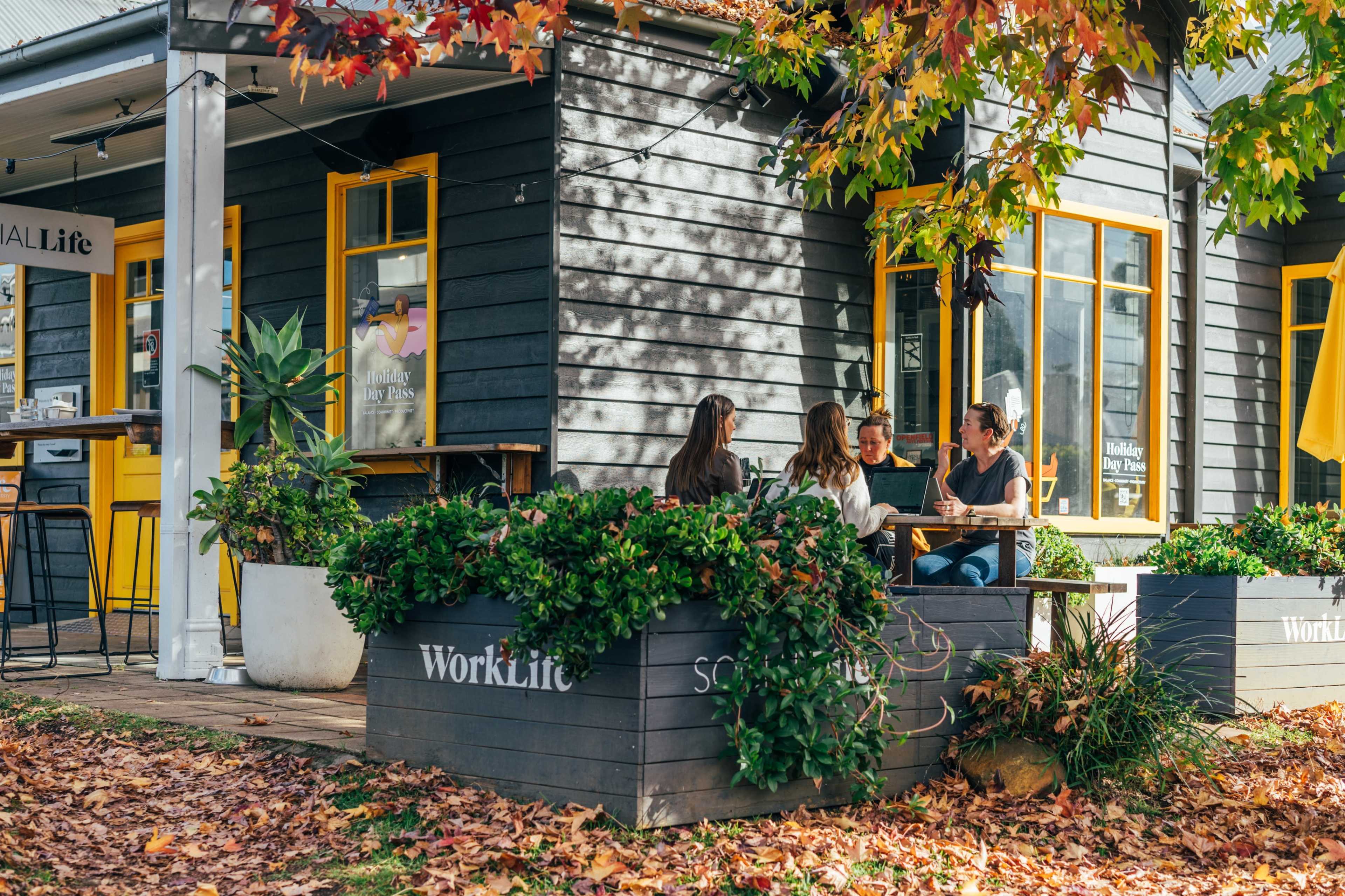 A group of four people is seated at a table in a cozy outdoor area surrounded by colorful autumn leaves and potted plants near a building with yellow accents.