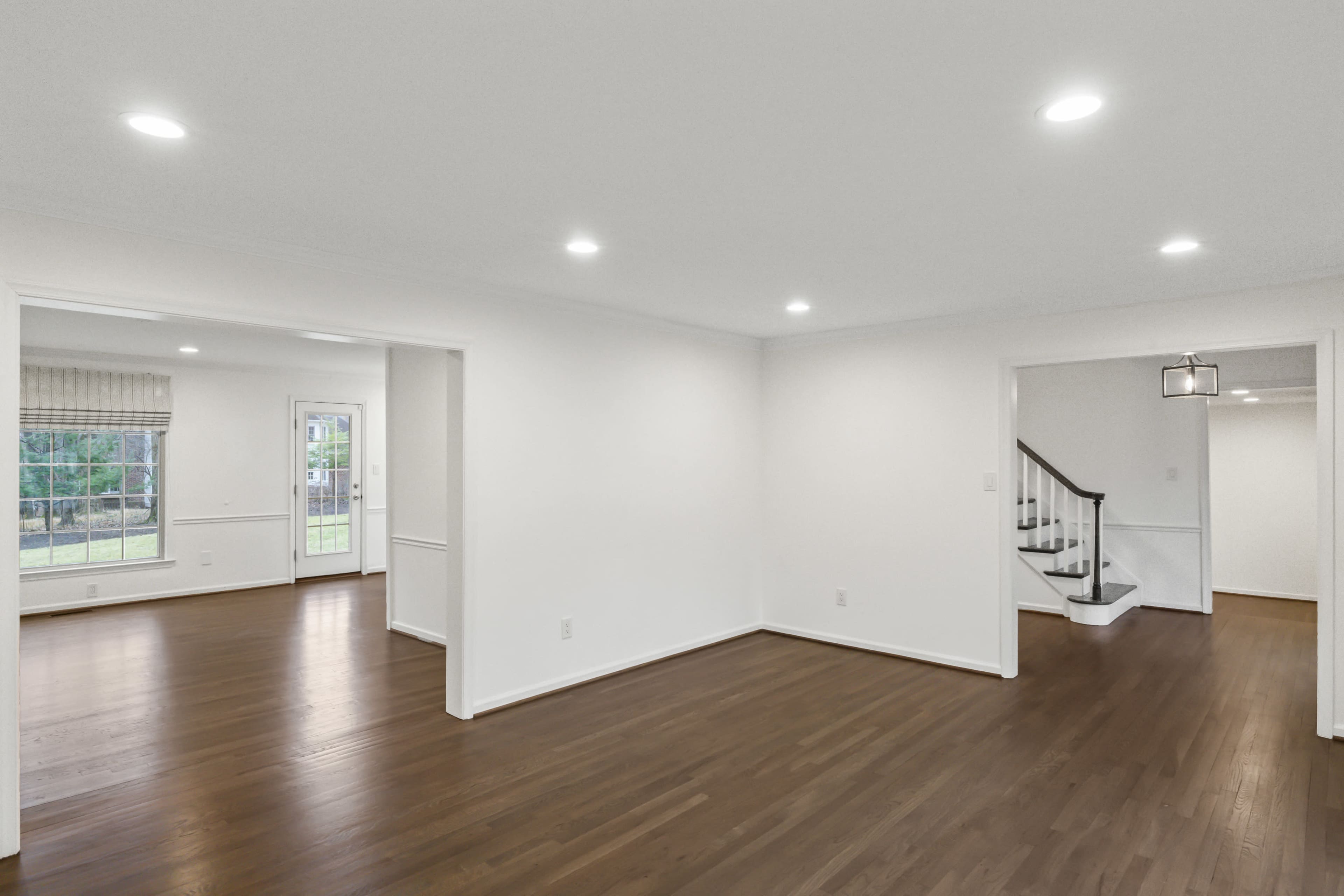 The image shows a spacious, well-lit interior of a home featuring hardwood floors, white walls, and an open layout connecting a sitting area and a staircase.