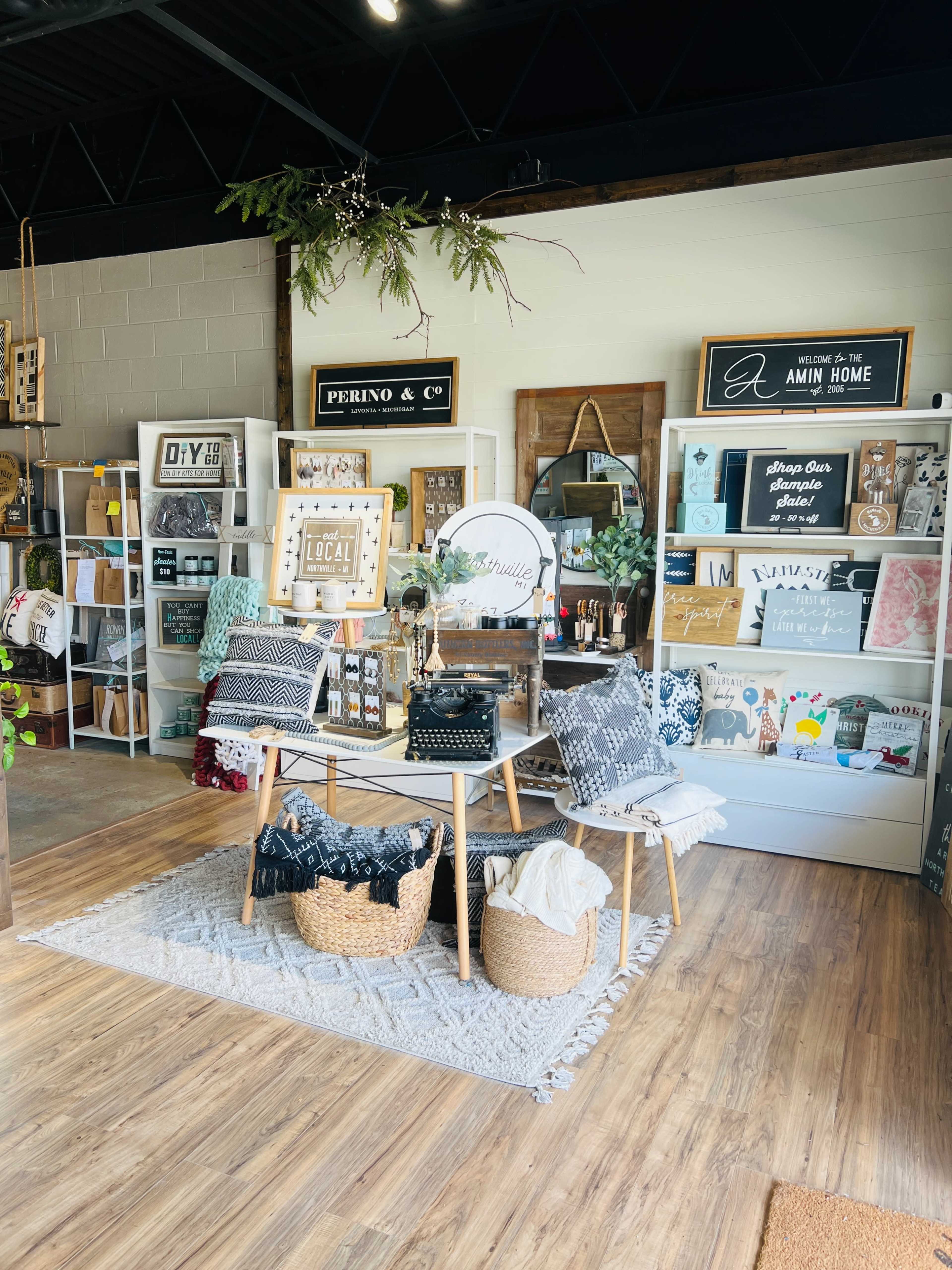 A well-organized retail space featuring shelves with home decor items, baskets, and decorative pillows arranged around a central table.