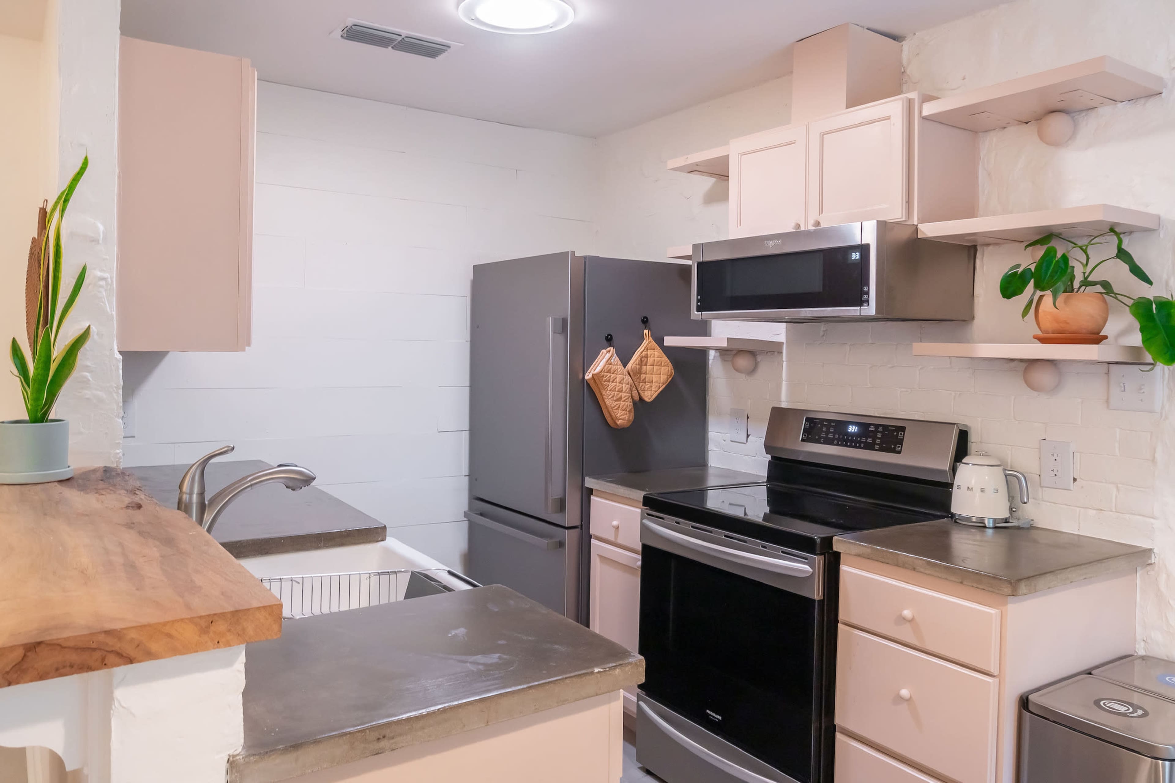 The image shows a modern kitchen with light-colored cabinets, a stainless steel refrigerator, and a black stove, featuring wooden shelves with plants and kitchenware.