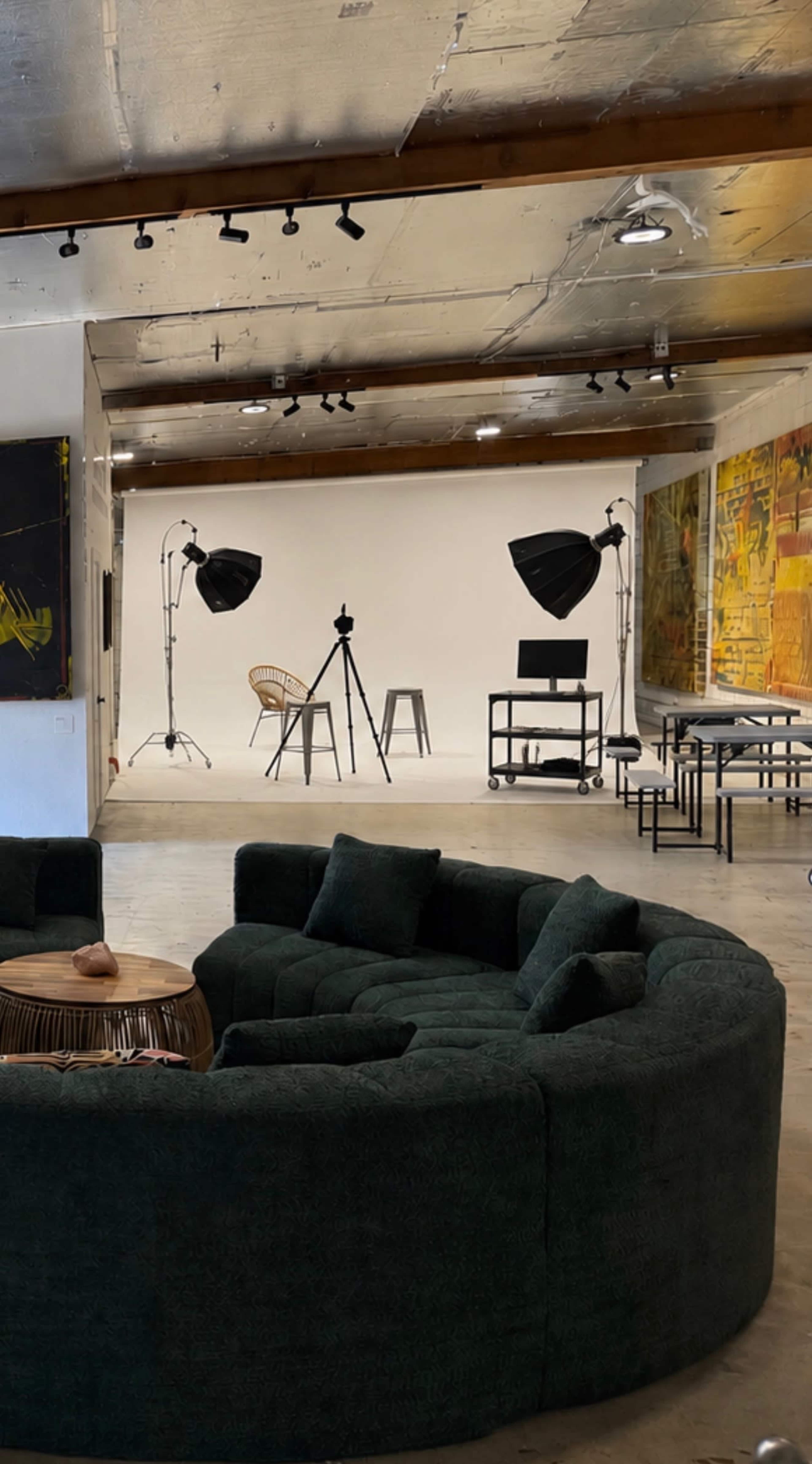 The image shows a spacious studio with a circular dark green sofa, a wooden coffee table, and various photography equipment set up against a plain white backdrop.