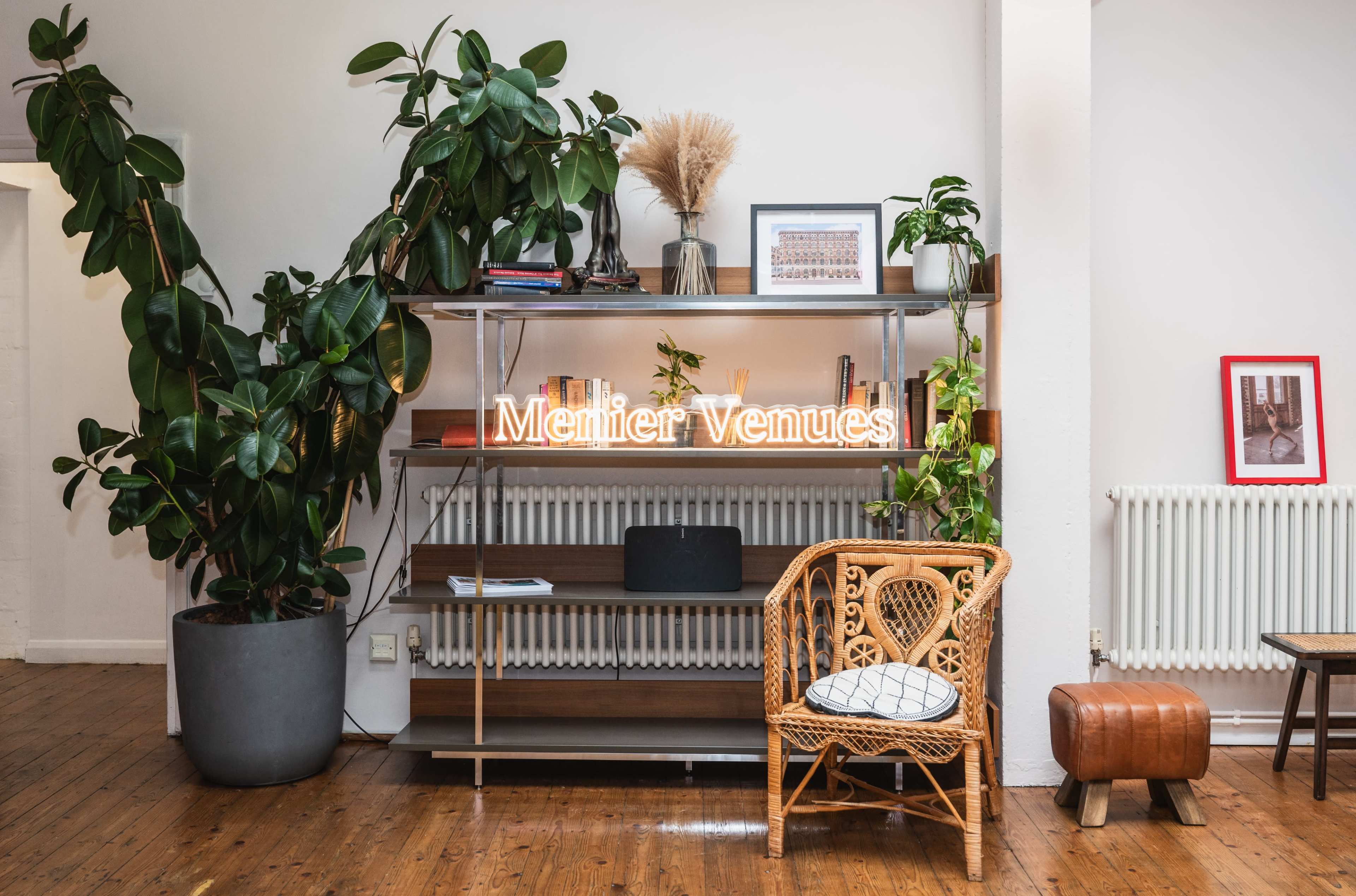 A modern interior features a wooden shelf with decorative plants, a glowing sign that reads "Merlier Venues," and a rattan chair next to a radiator.