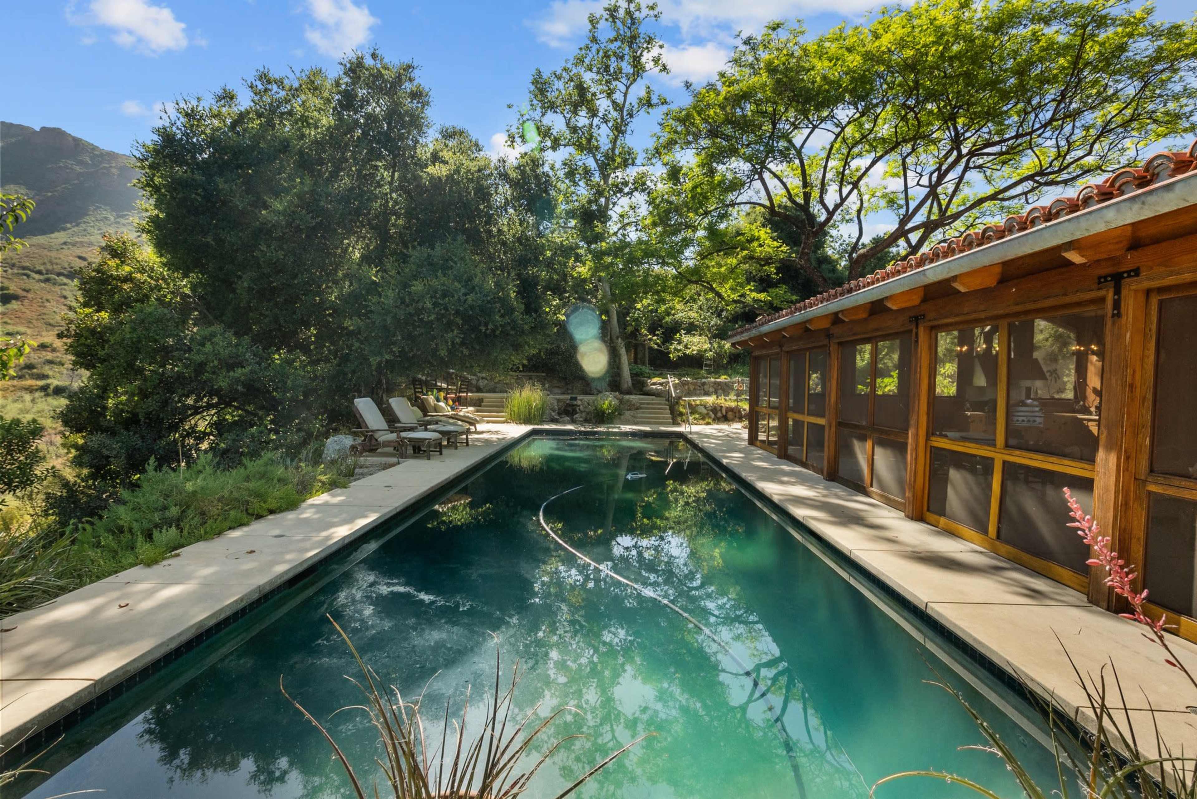 The image shows a serene outdoor pool surrounded by lush greenery and a wooden structure with large windows adjacent to it.