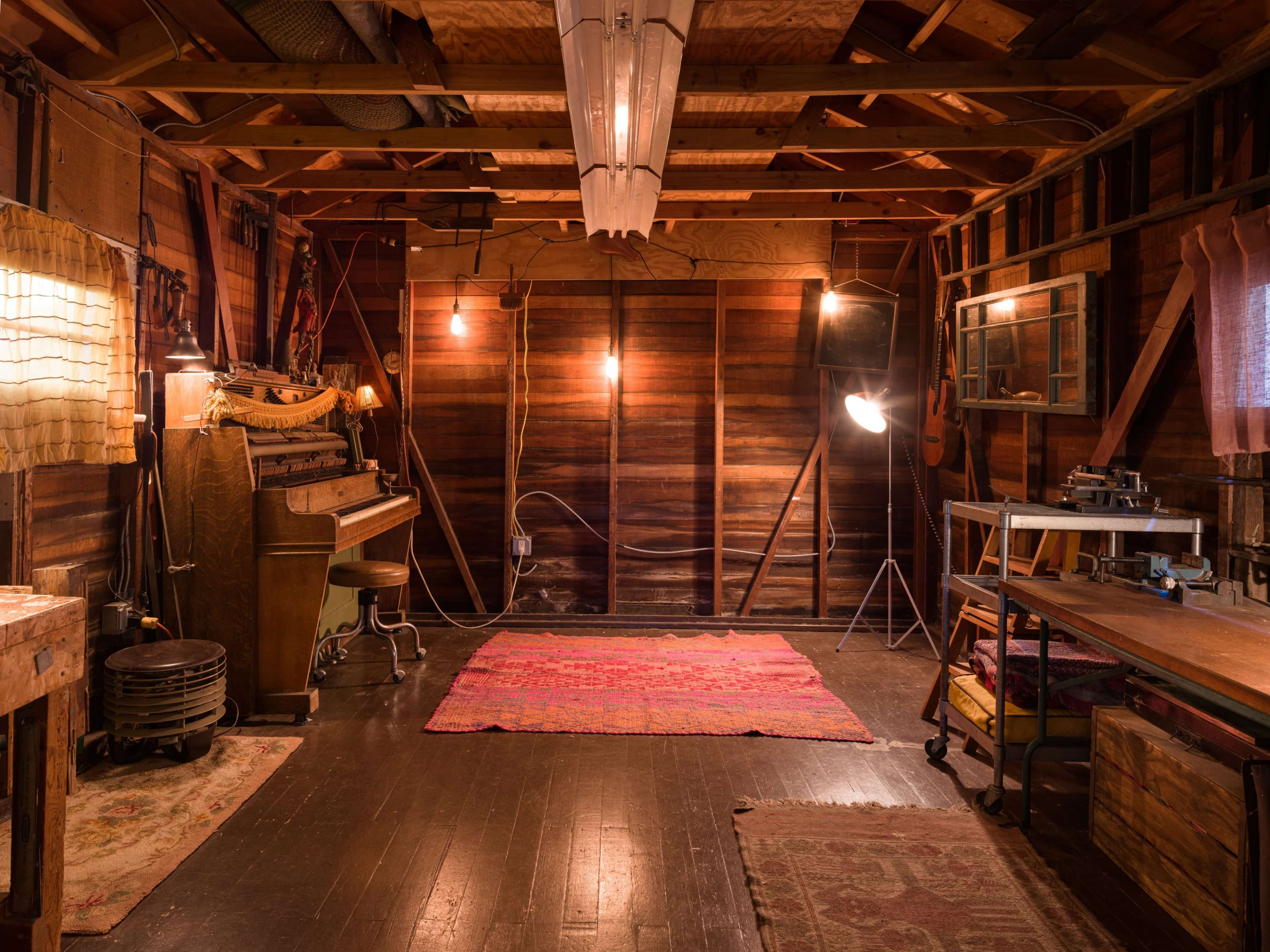 The image shows a wooden interior space featuring a vintage piano, a patterned rug, various lighting fixtures, and a work table.