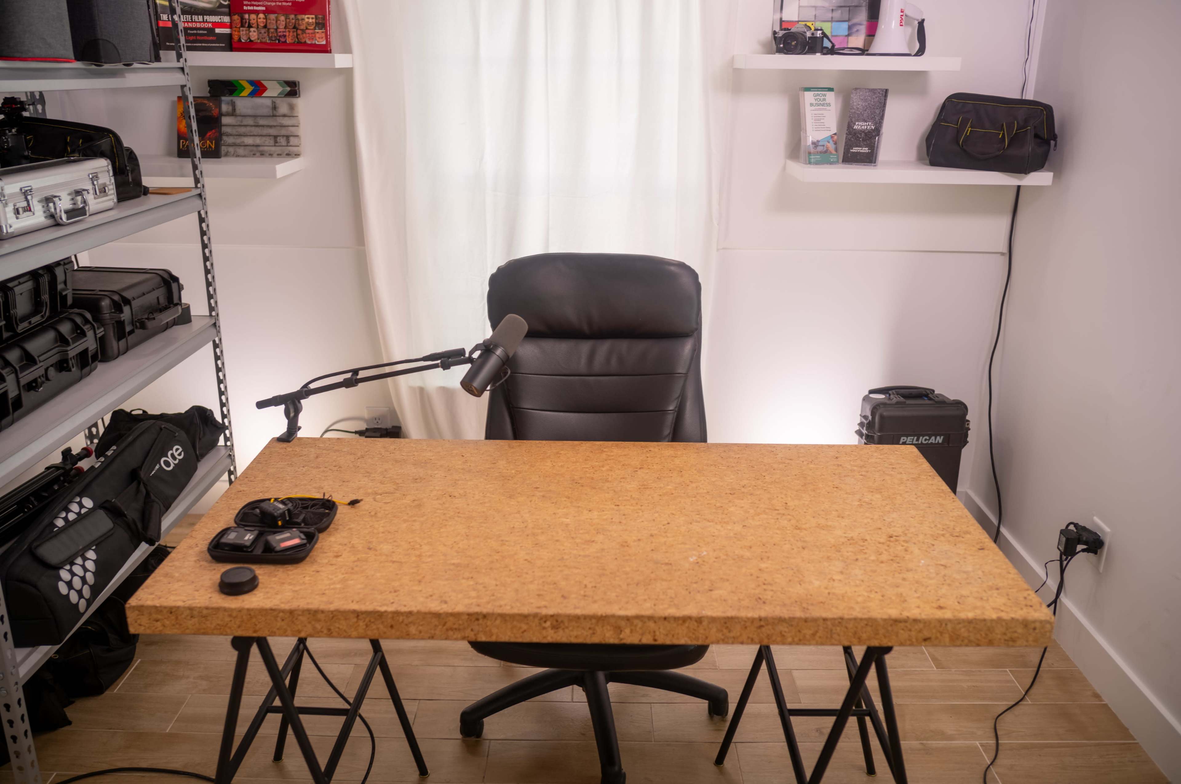 A black office chair is positioned in front of a wooden table in a well-lit room with shelves displaying various items.