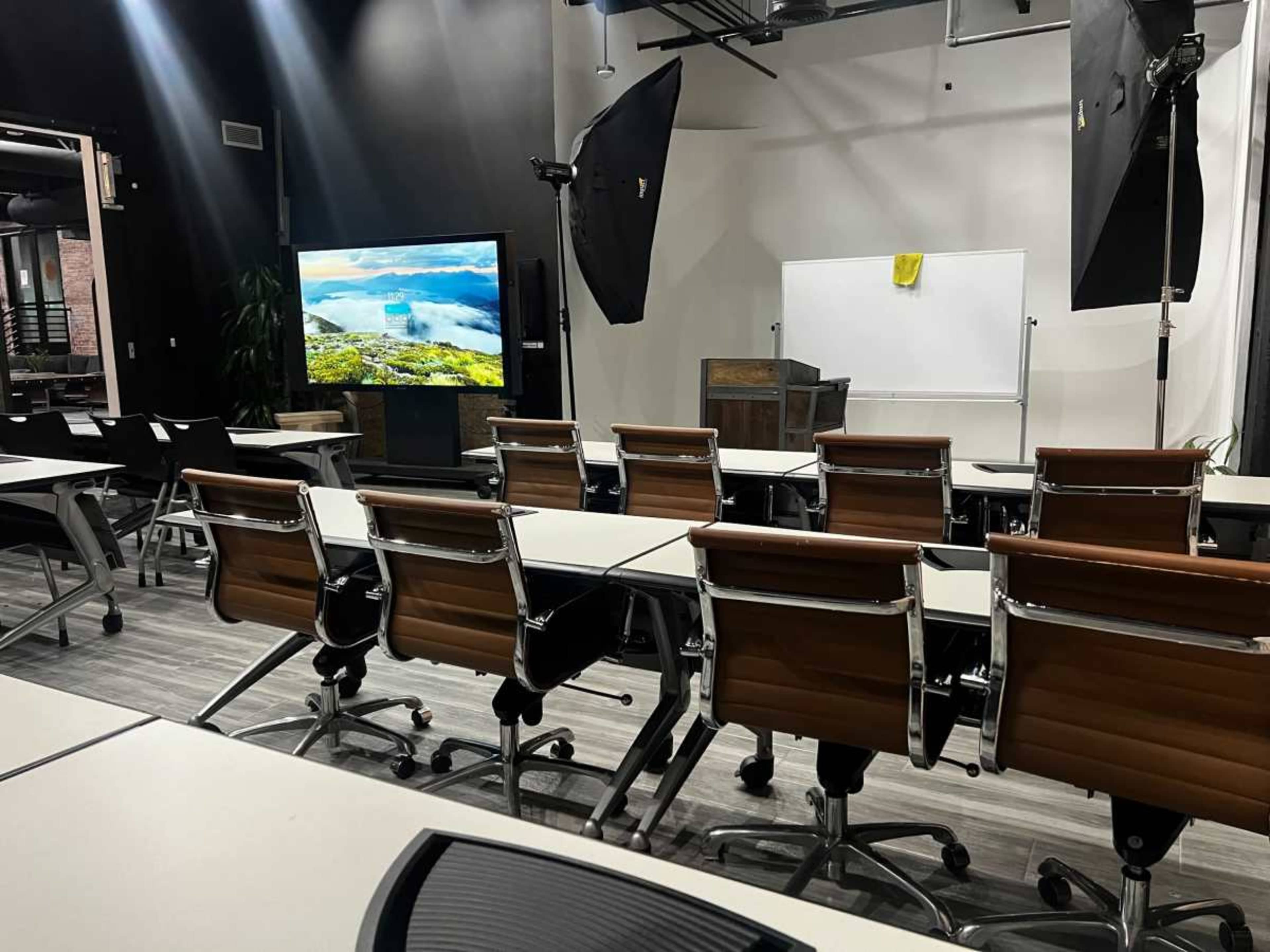 A modern classroom setup features rows of chairs and tables facing a large screen, a whiteboard, and studio lighting equipment.