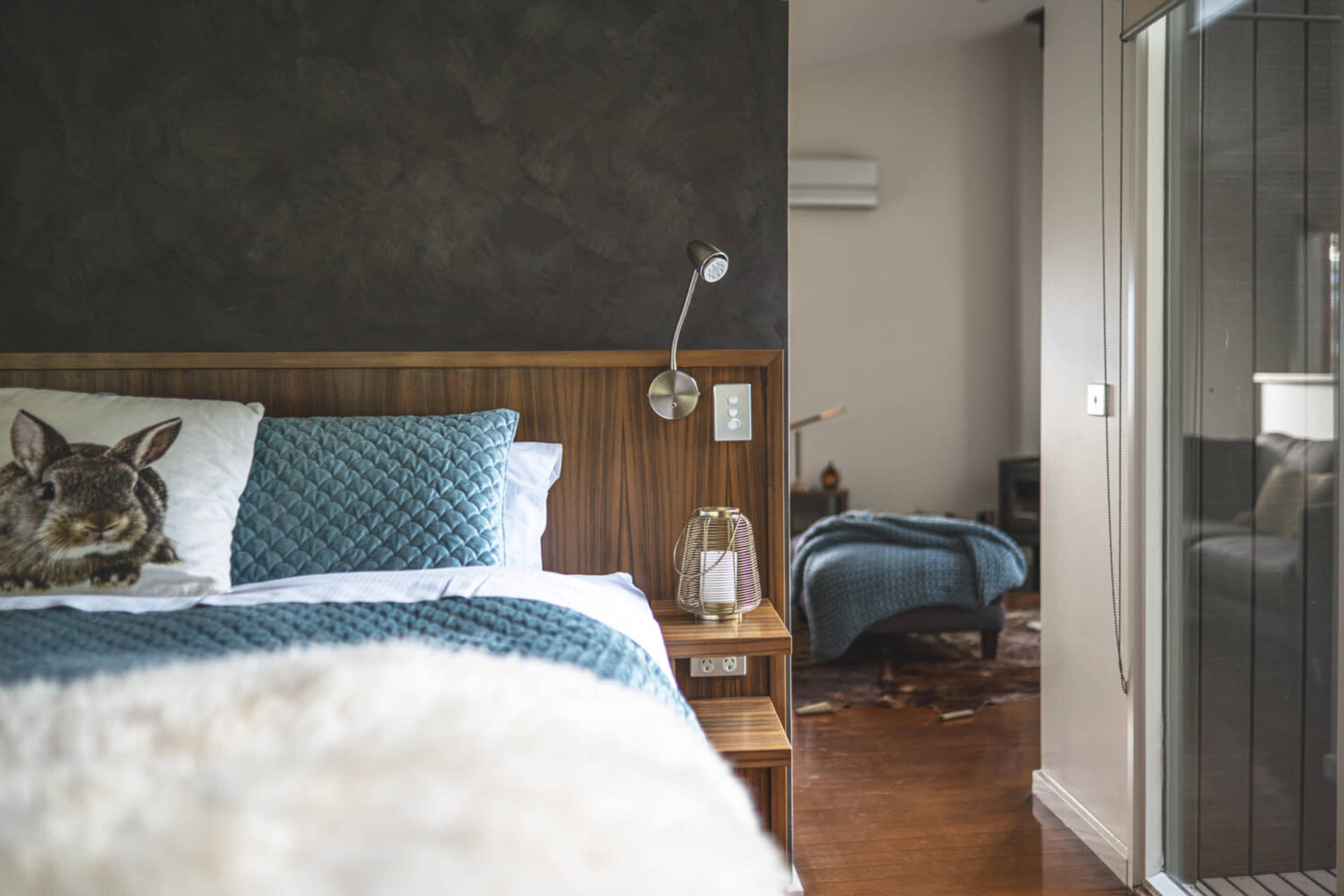 A cozy bedroom with a wooden bed featuring a quilted blue blanket, a bedside table with a lamp, and a visible second bed in the background.
