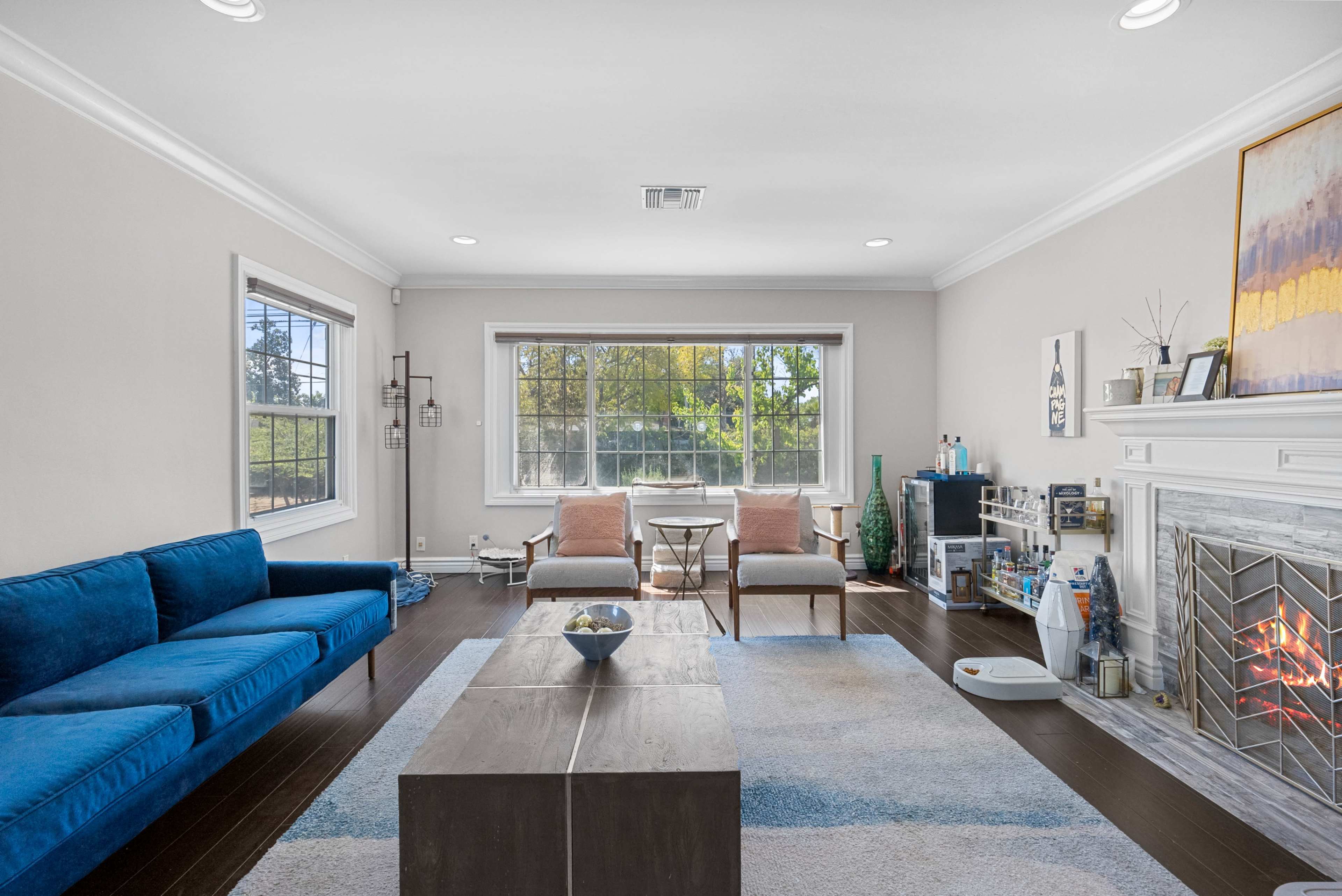 A living room features a blue sofa, two chairs, a coffee table, and a fireplace with a large window providing natural light.