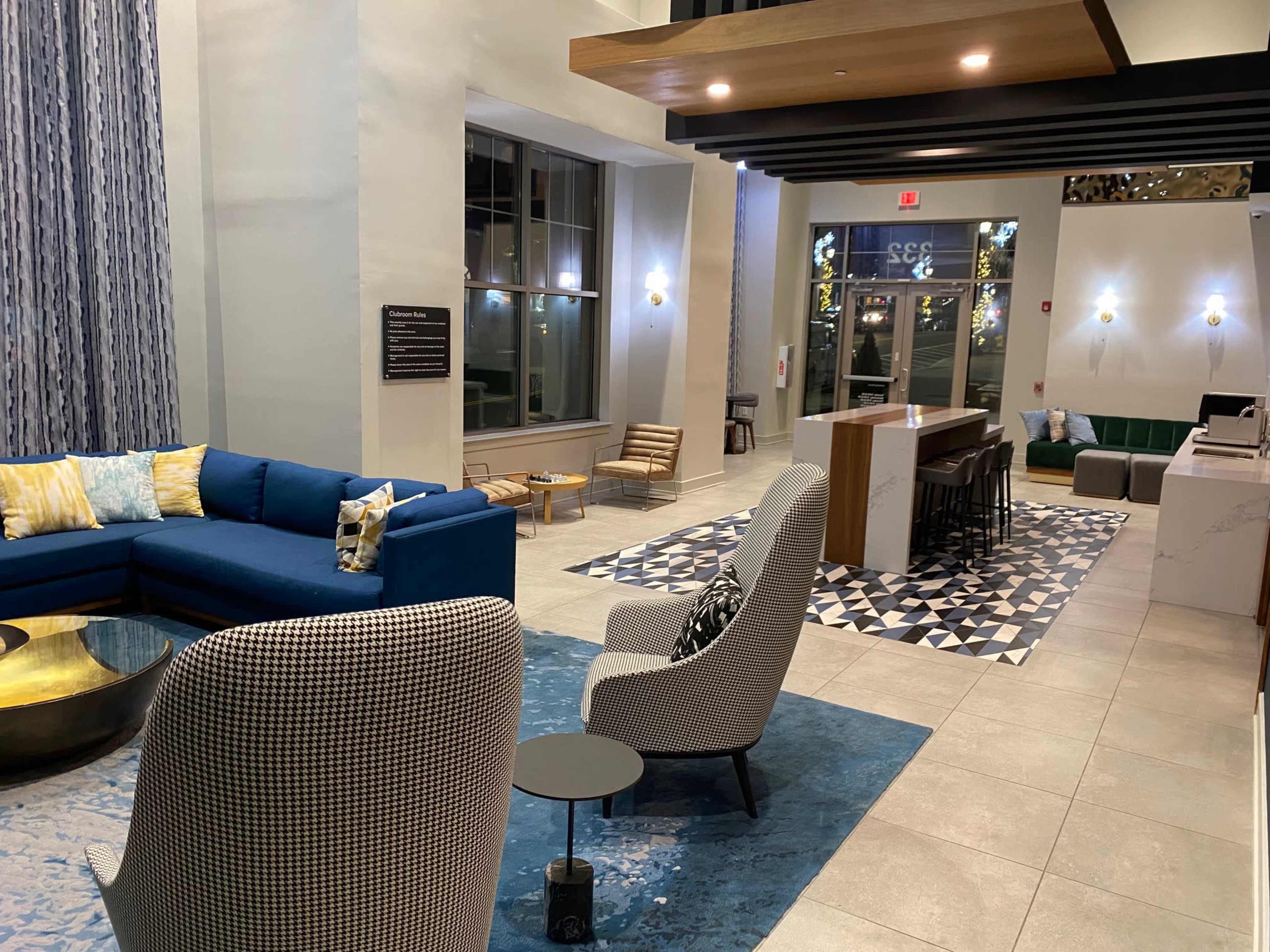 An open lobby area features a blue sectional sofa, patterned armchairs, a round coffee table, and a checkered rug, with a reception desk in the background.