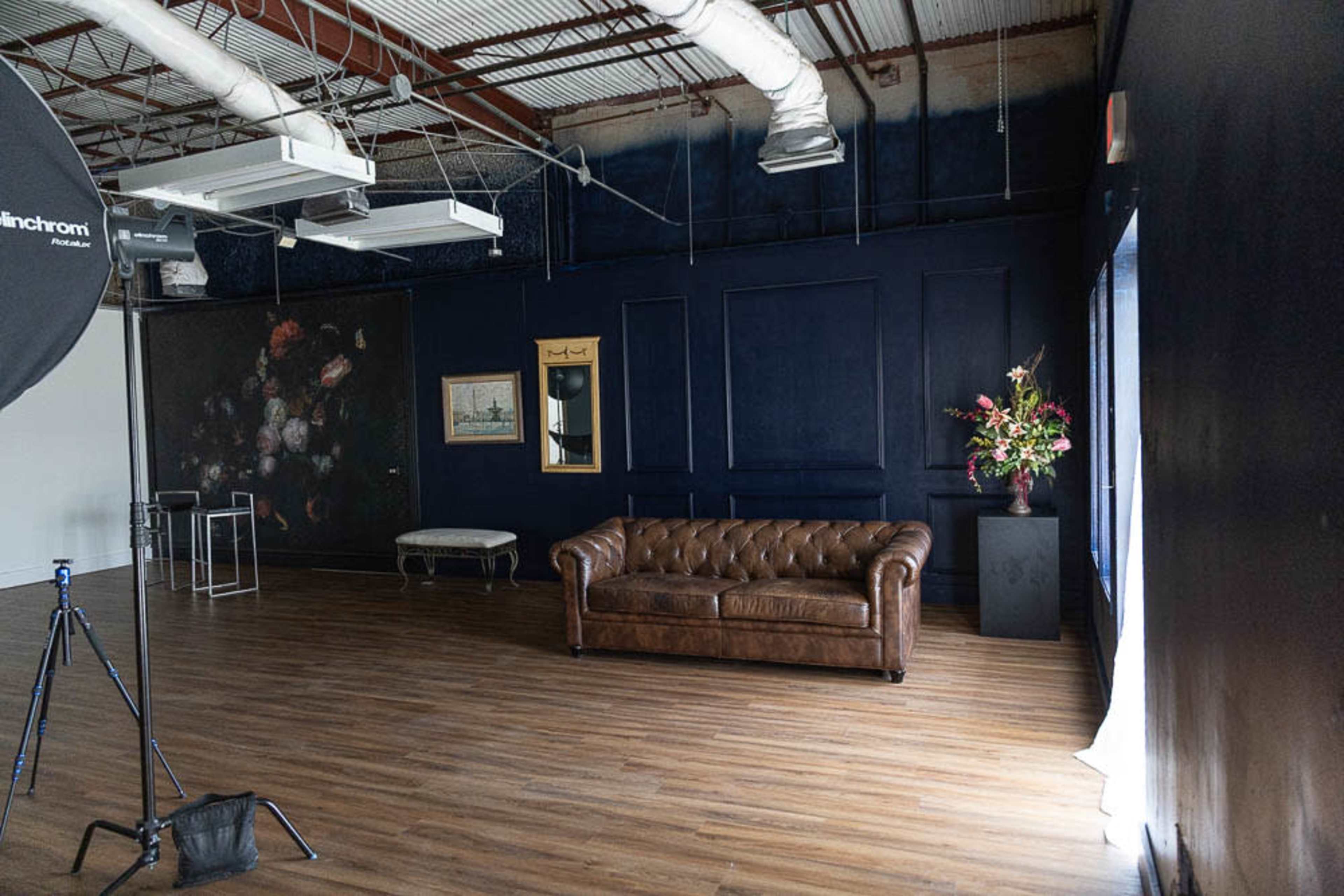 The image shows a spacious studio interior featuring a brown leather chesterfield sofa, a floral arrangement in a vase, and a dark blue wall with artwork.