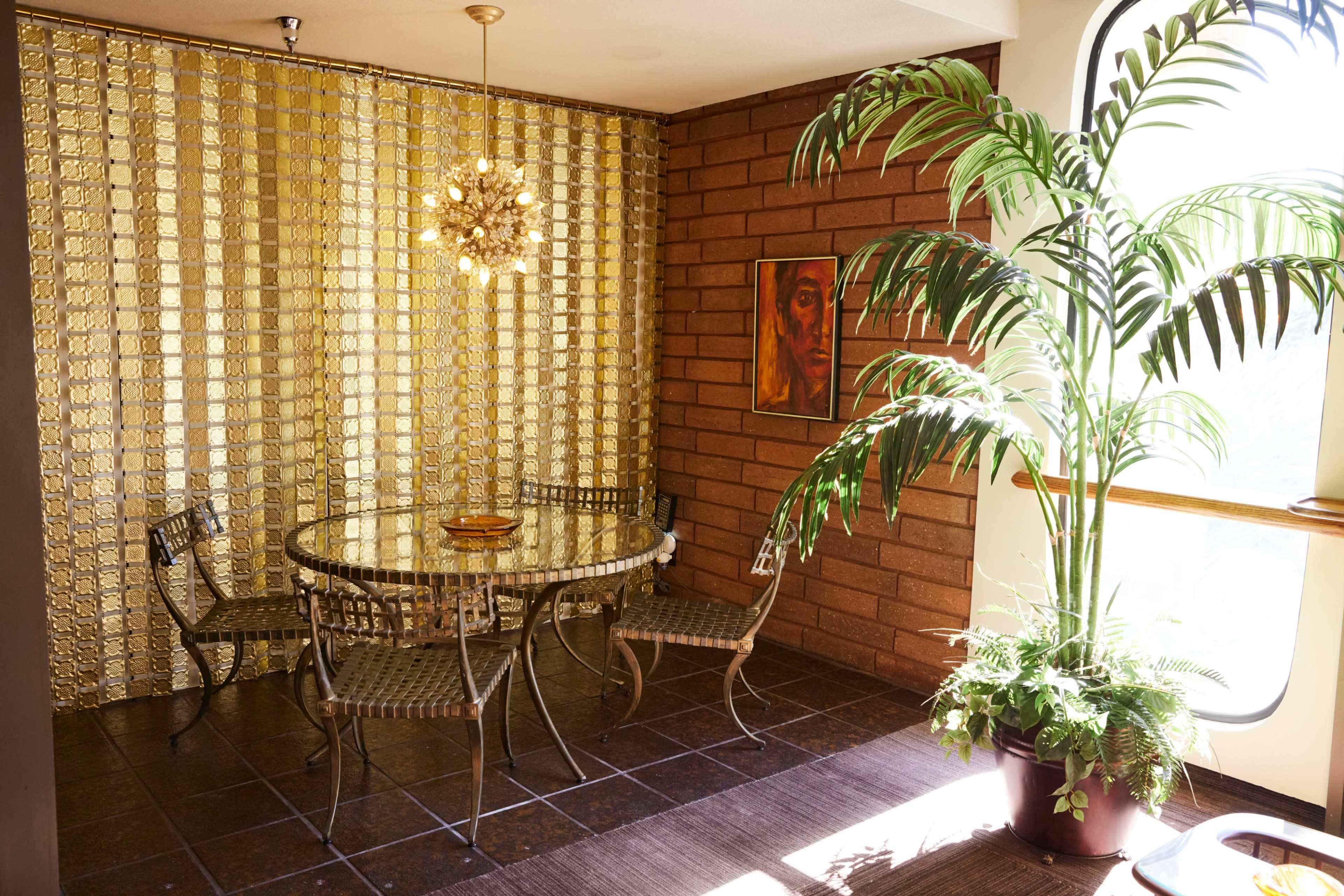 A dining area features a circular glass table surrounded by four metal chairs, with a golden wall and a large potted plant beside a window.