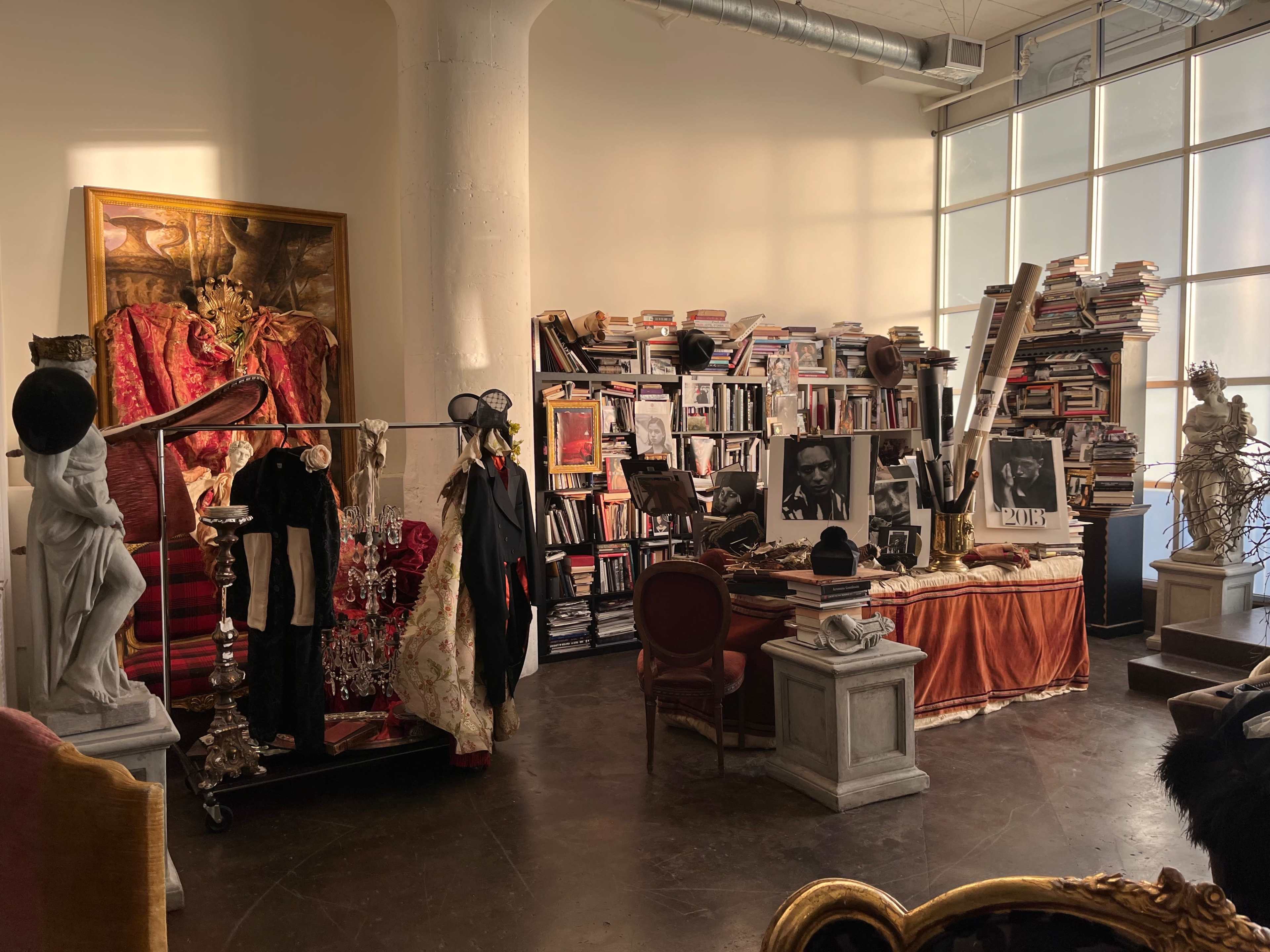 A spacious room filled with bookshelves, clothing on display, and various artistic decorations, including a large painting and sculptures.