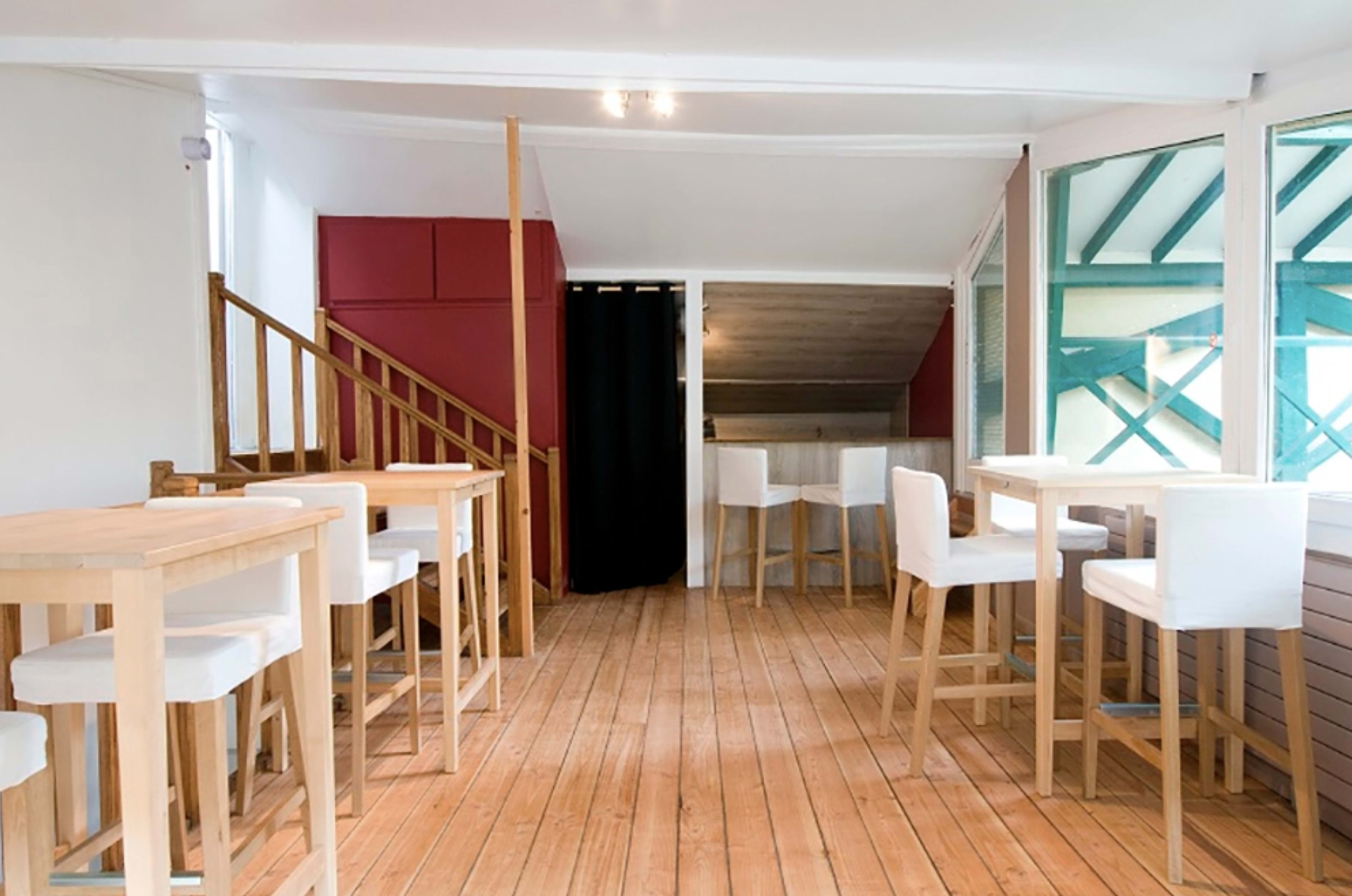 A bright interior of a wooden-floored café featuring several tall tables with white stools and a staircase leading to another level.