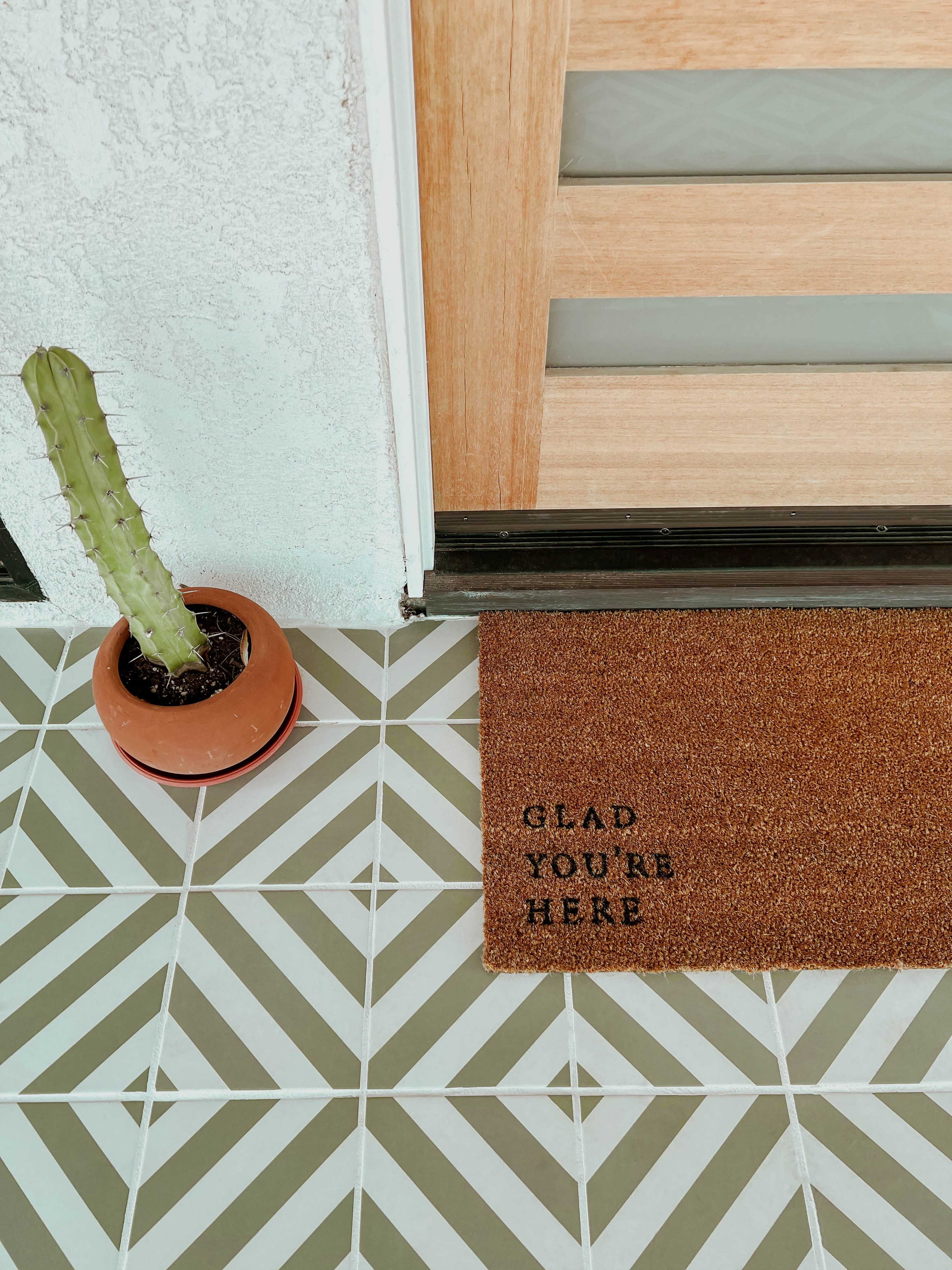 A welcome mat that reads "GLAD YOU'RE HERE" sits on patterned tile flooring next to a potted cactus.