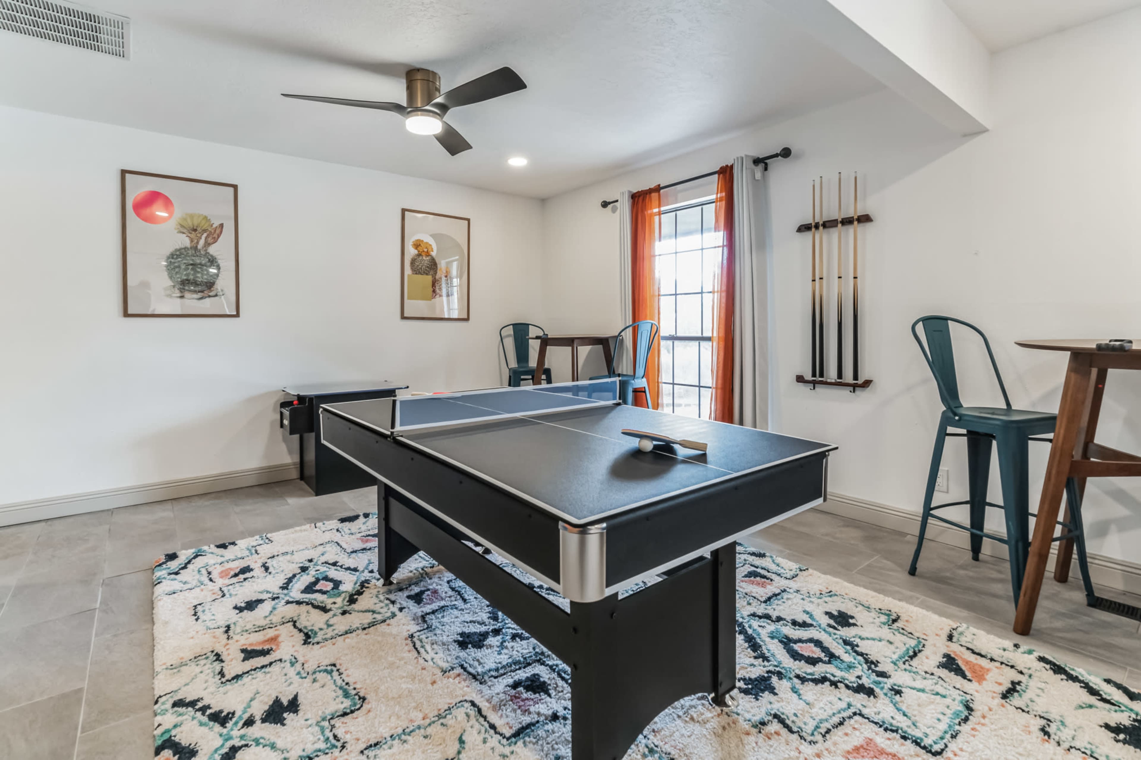 A game room features two ping pong tables, a small dining area, and a window with orange curtains.