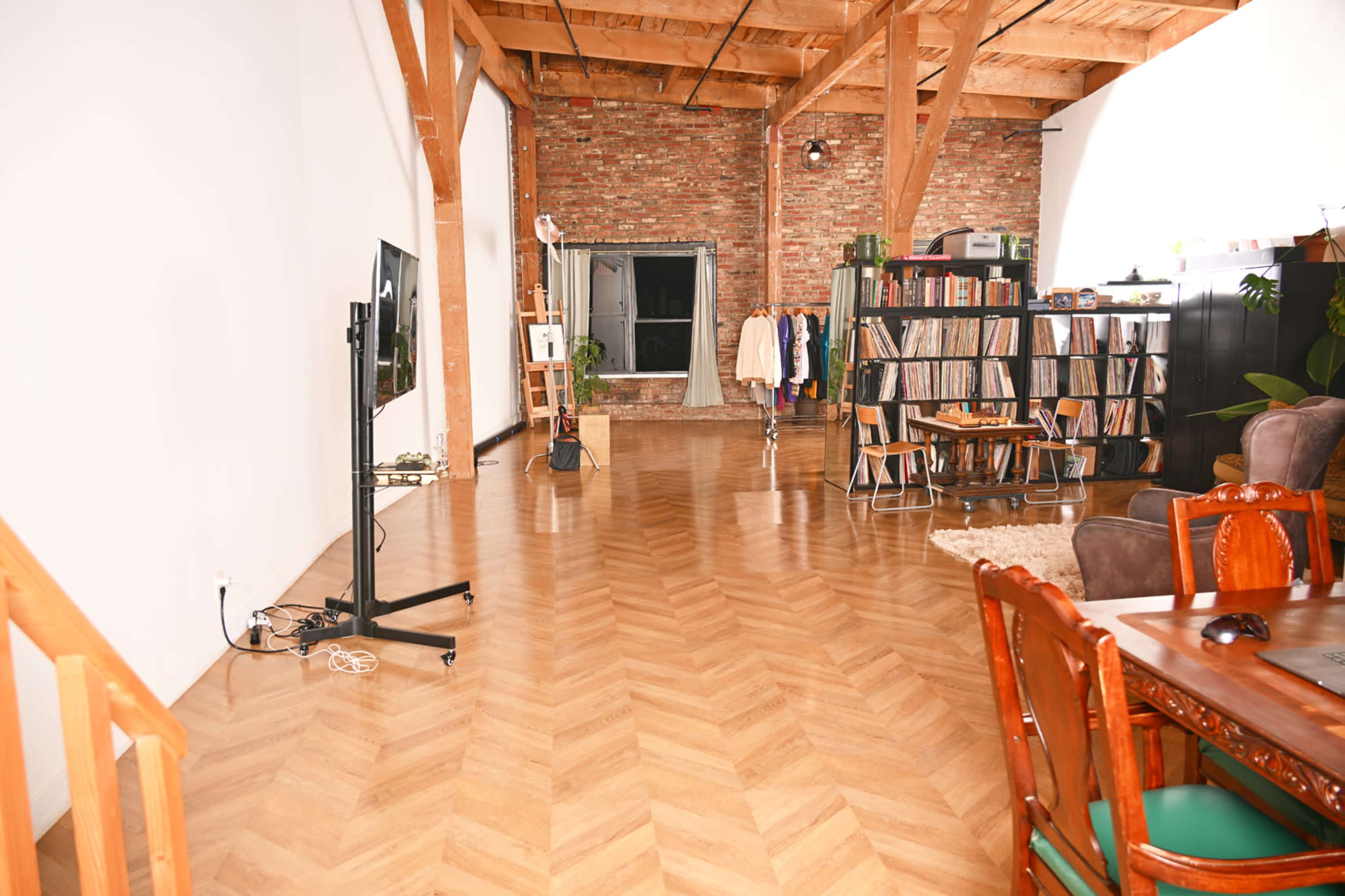 The image shows a spacious room with wooden flooring, exposed brick walls, and a mixture of furniture including a dining table, a TV stand, and a bookshelf.