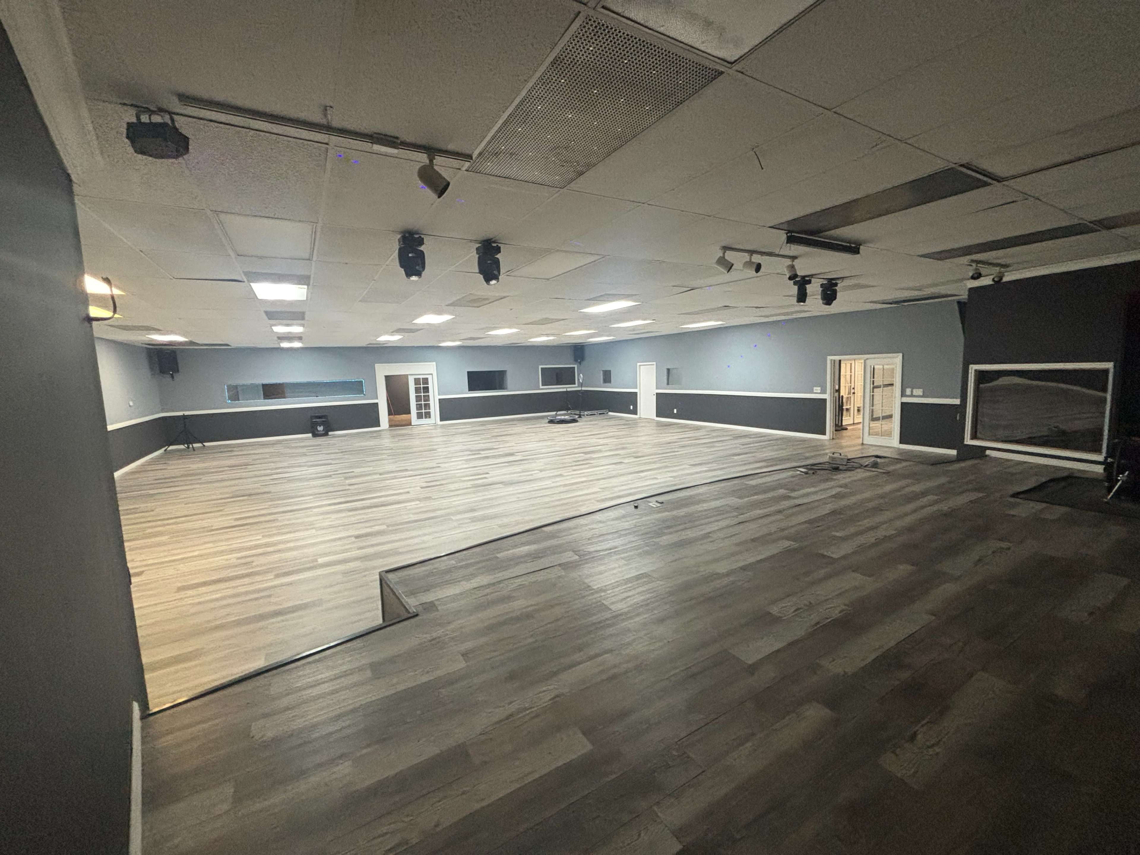 The image shows an empty, spacious interior room with wooden flooring and multiple recessed lights.