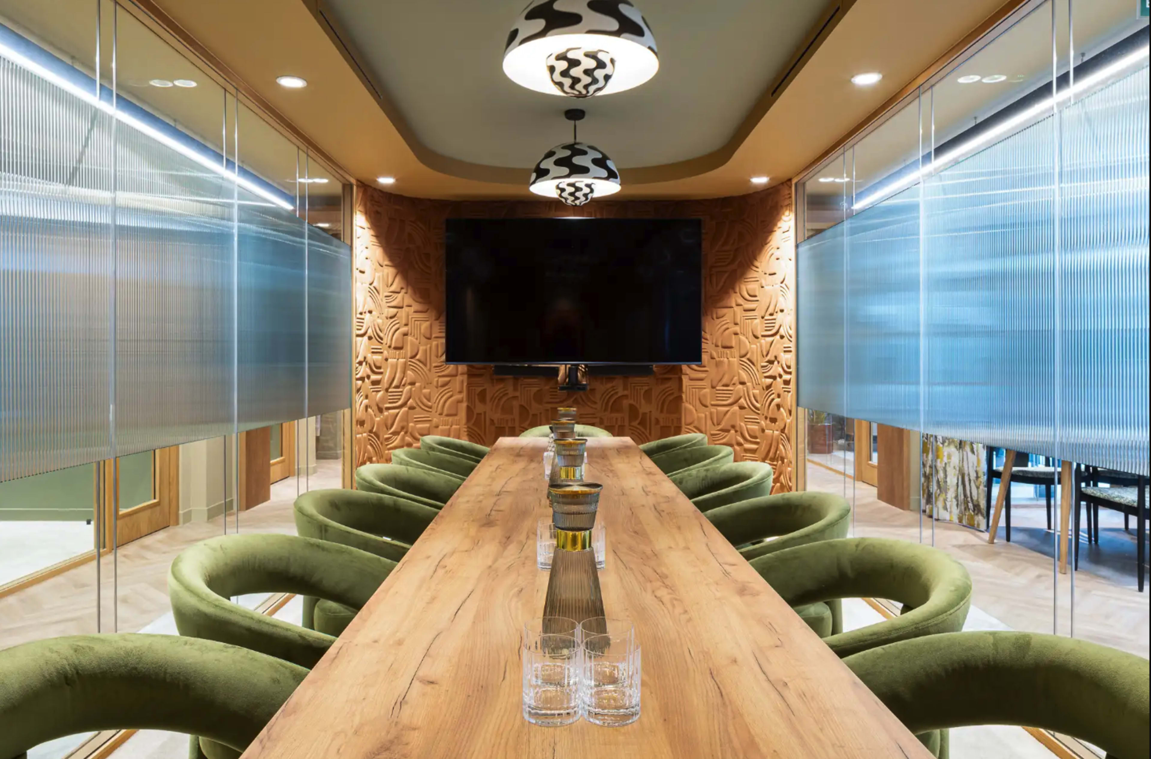 A modern conference room features a long wooden table surrounded by green chairs, with decorative walls and a large screen at one end.