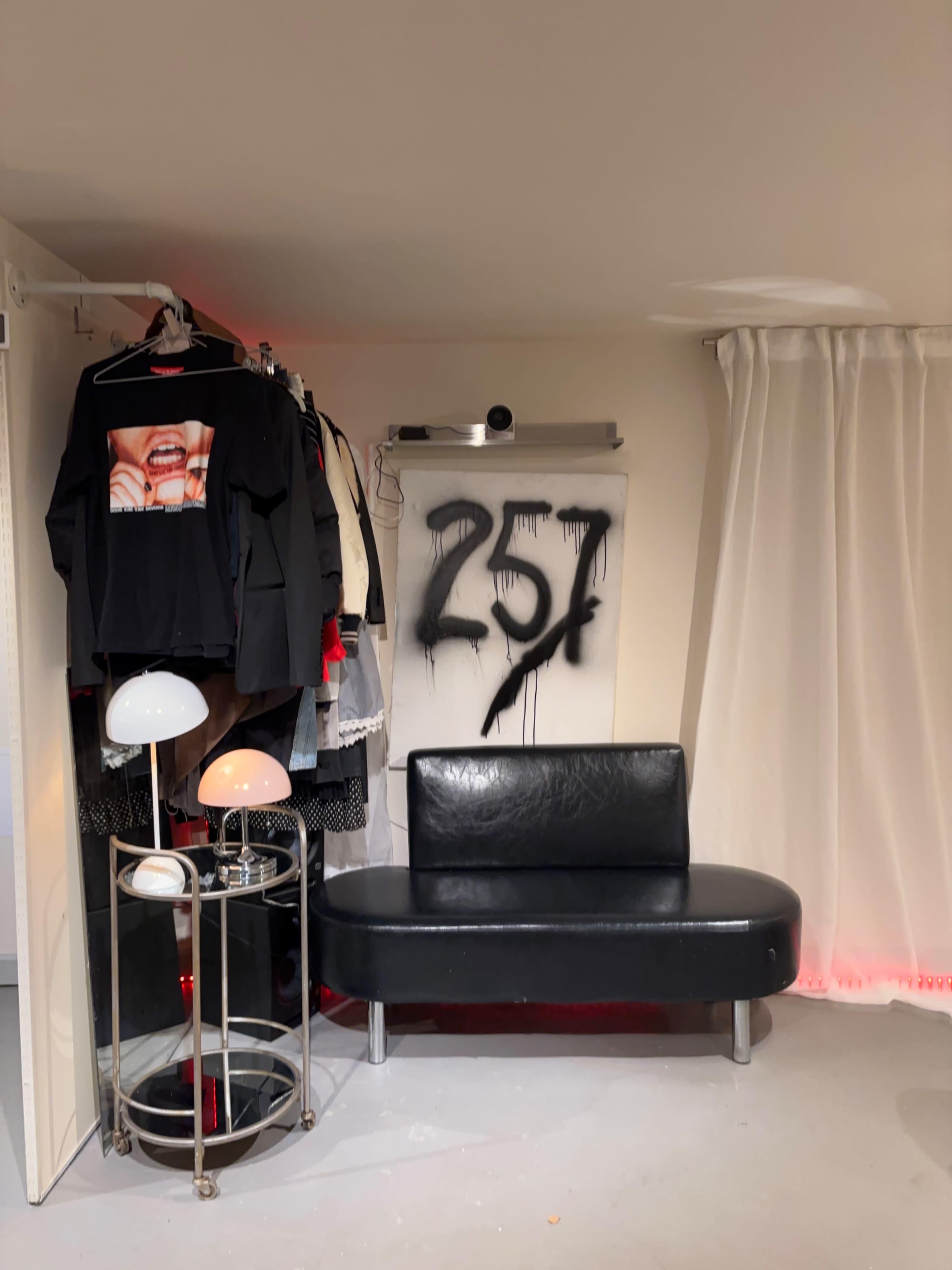 The image shows a small room featuring a black couch, a clothing rack with various garments, a lamp, and a wall with the number "257" painted on it.