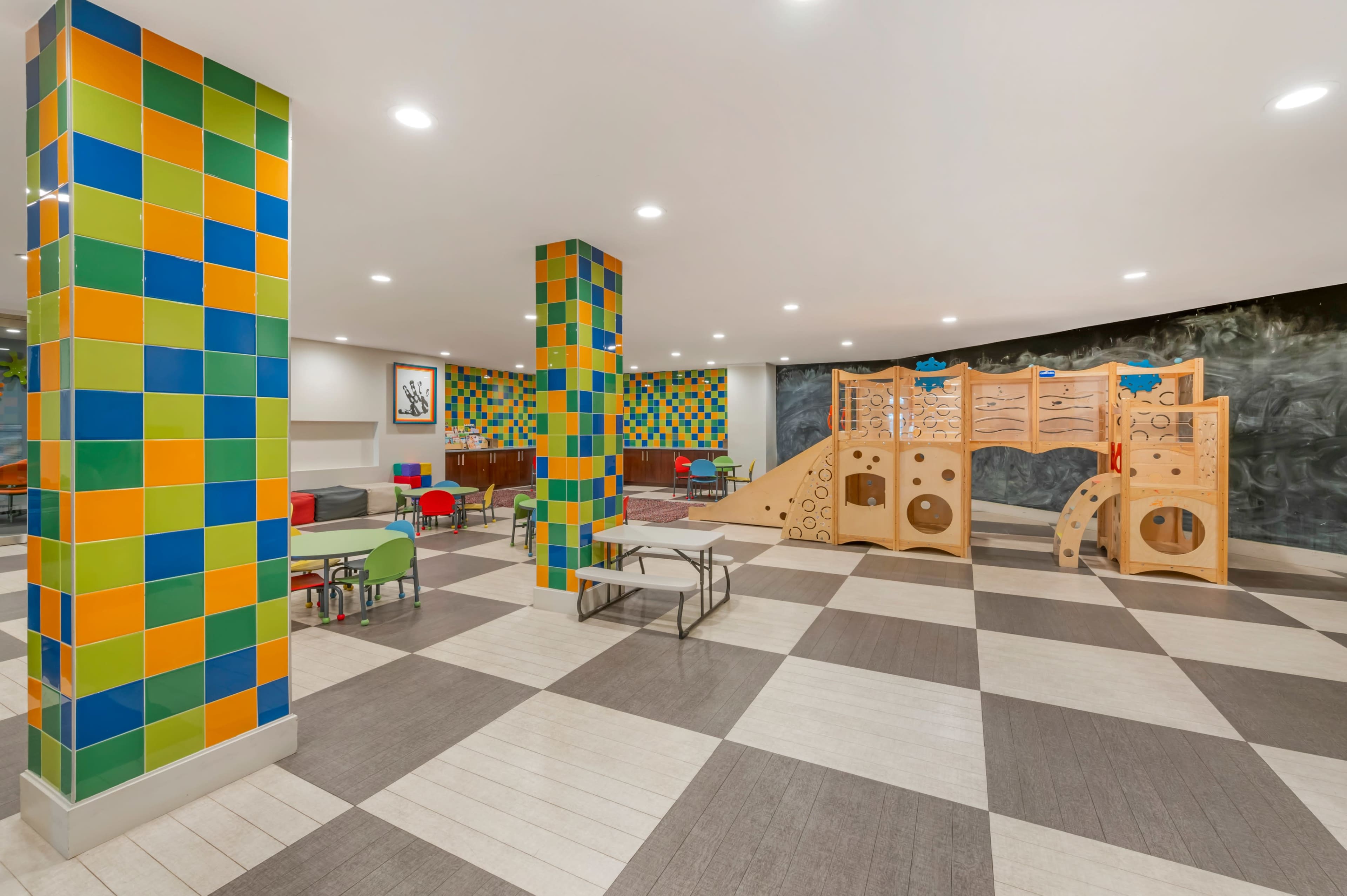 The image shows a brightly colored play area featuring colorful tiled columns, a wooden play structure, and various seating arrangements on a checkered floor.