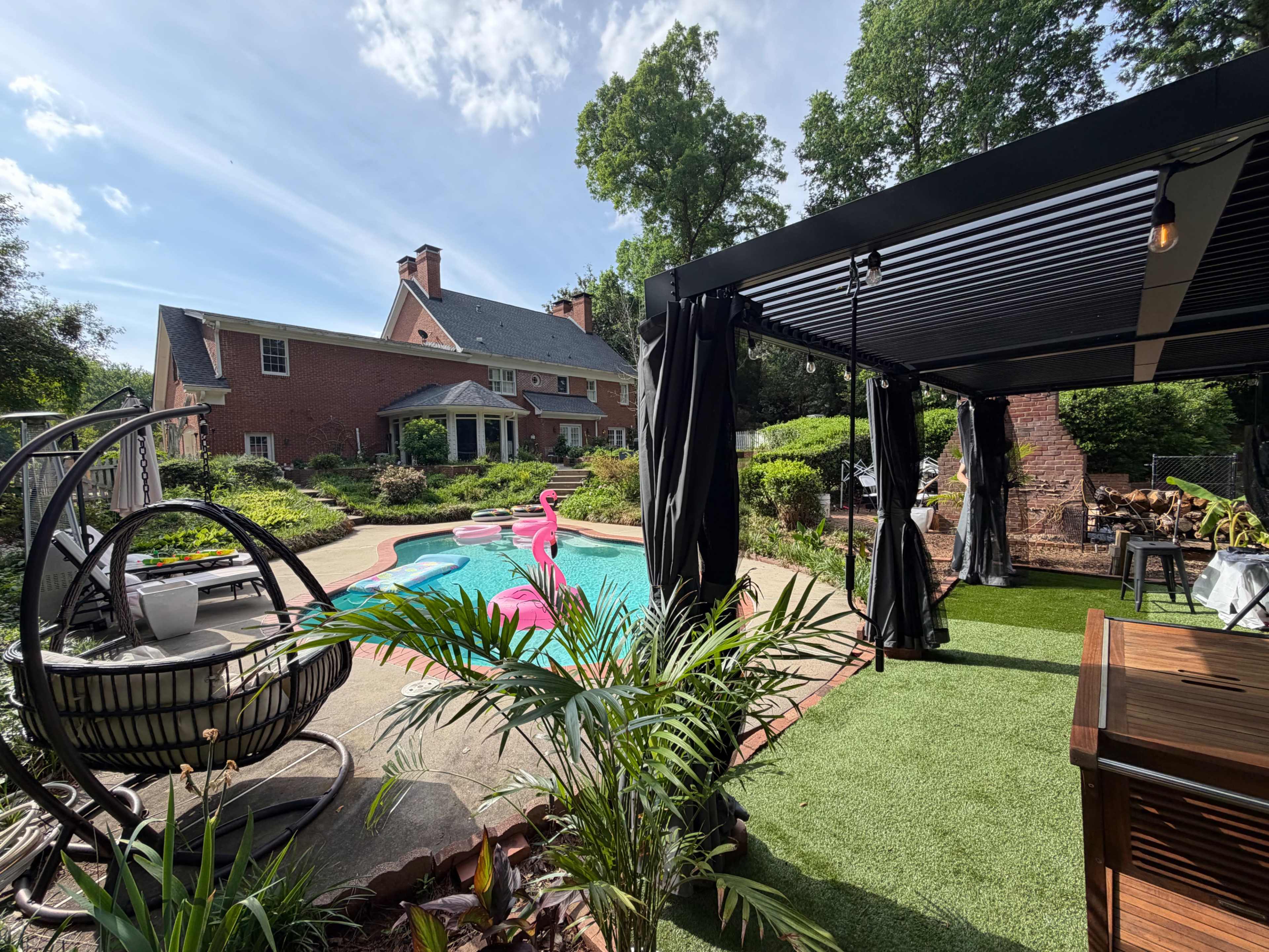 The image shows a backyard with a swimming pool, a pink flamingo float, a gazebo-like structure, and a well-kept garden in front of a large brick house.