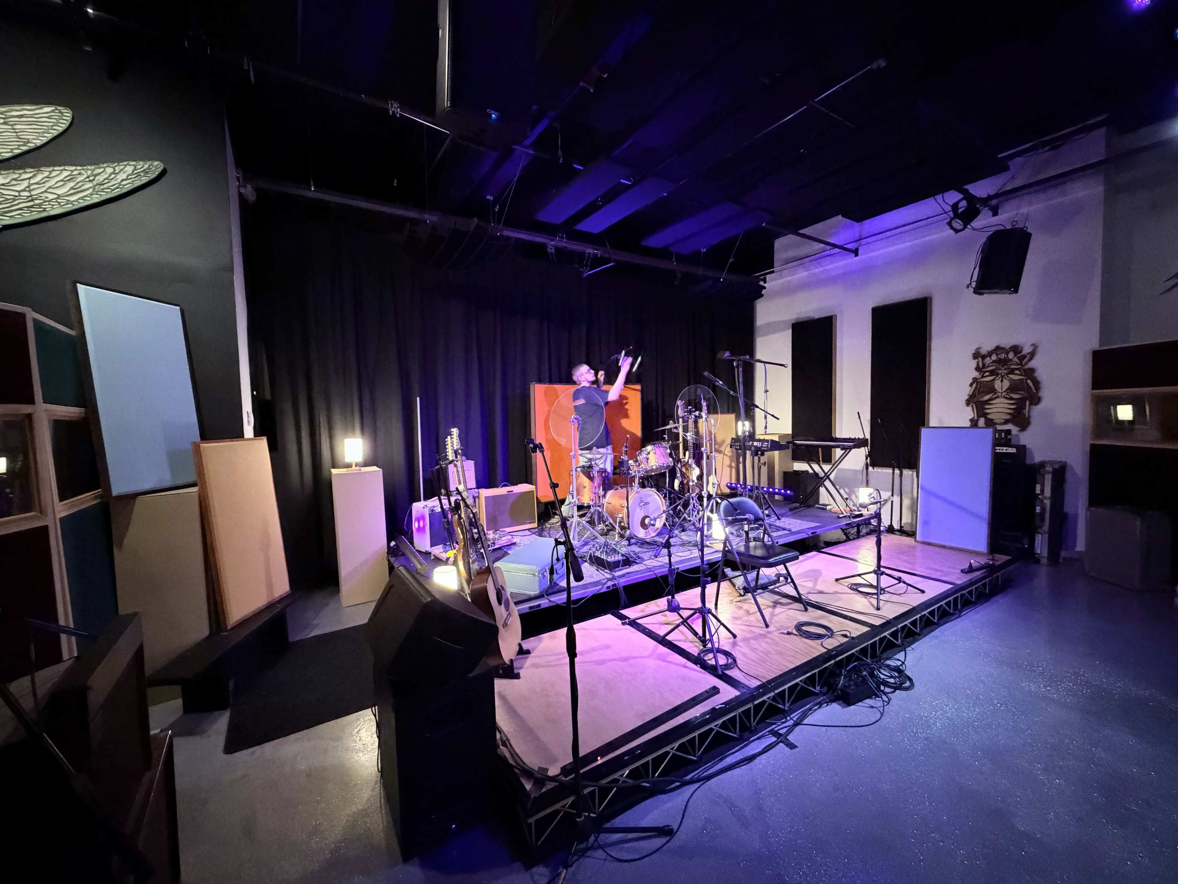 A musician stands on stage in a dimly lit room surrounded by various musical instruments and equipment.