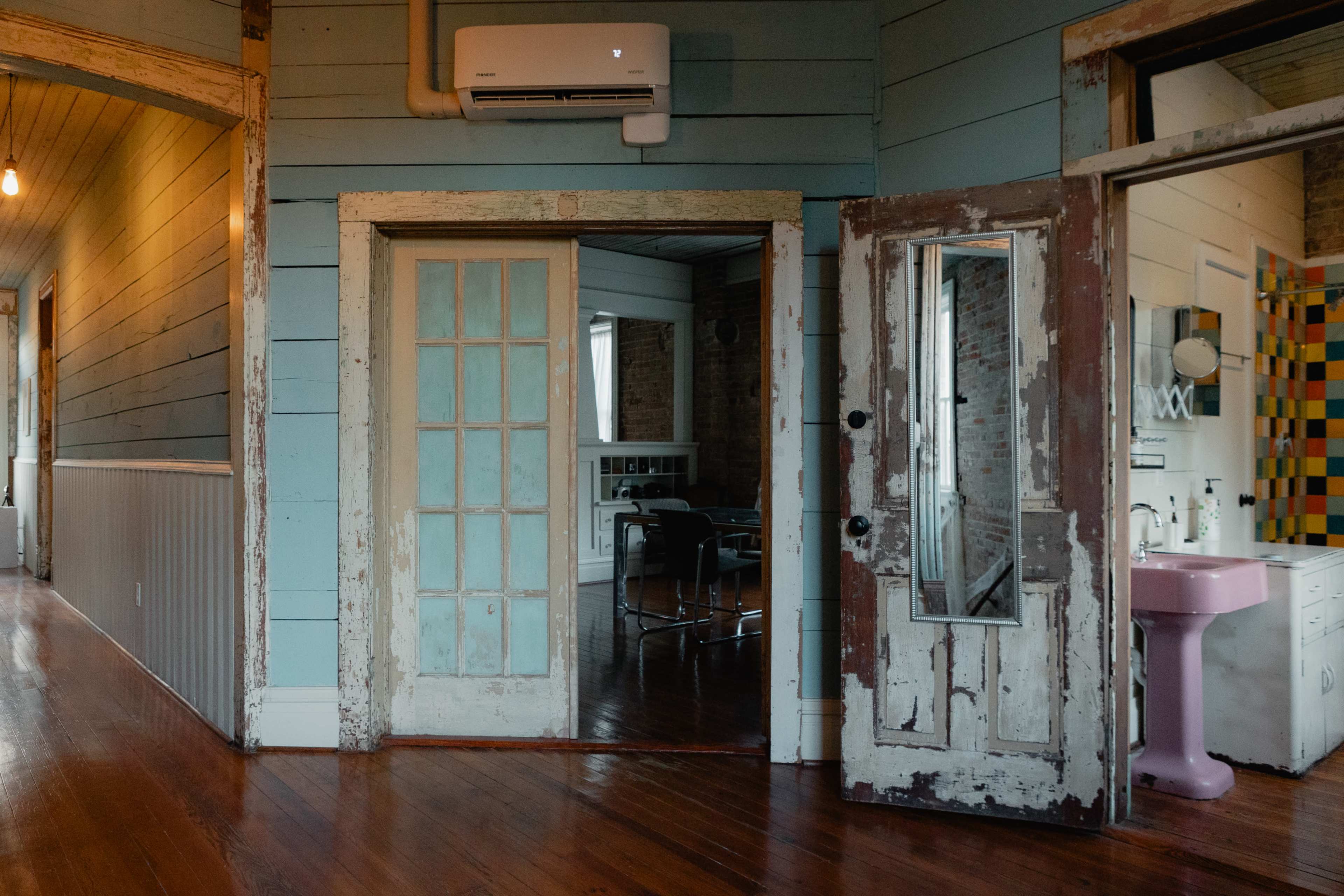 The image shows a room with rustic wooden walls, vintage doors, and a pink sink visible in an adjoining bathroom.