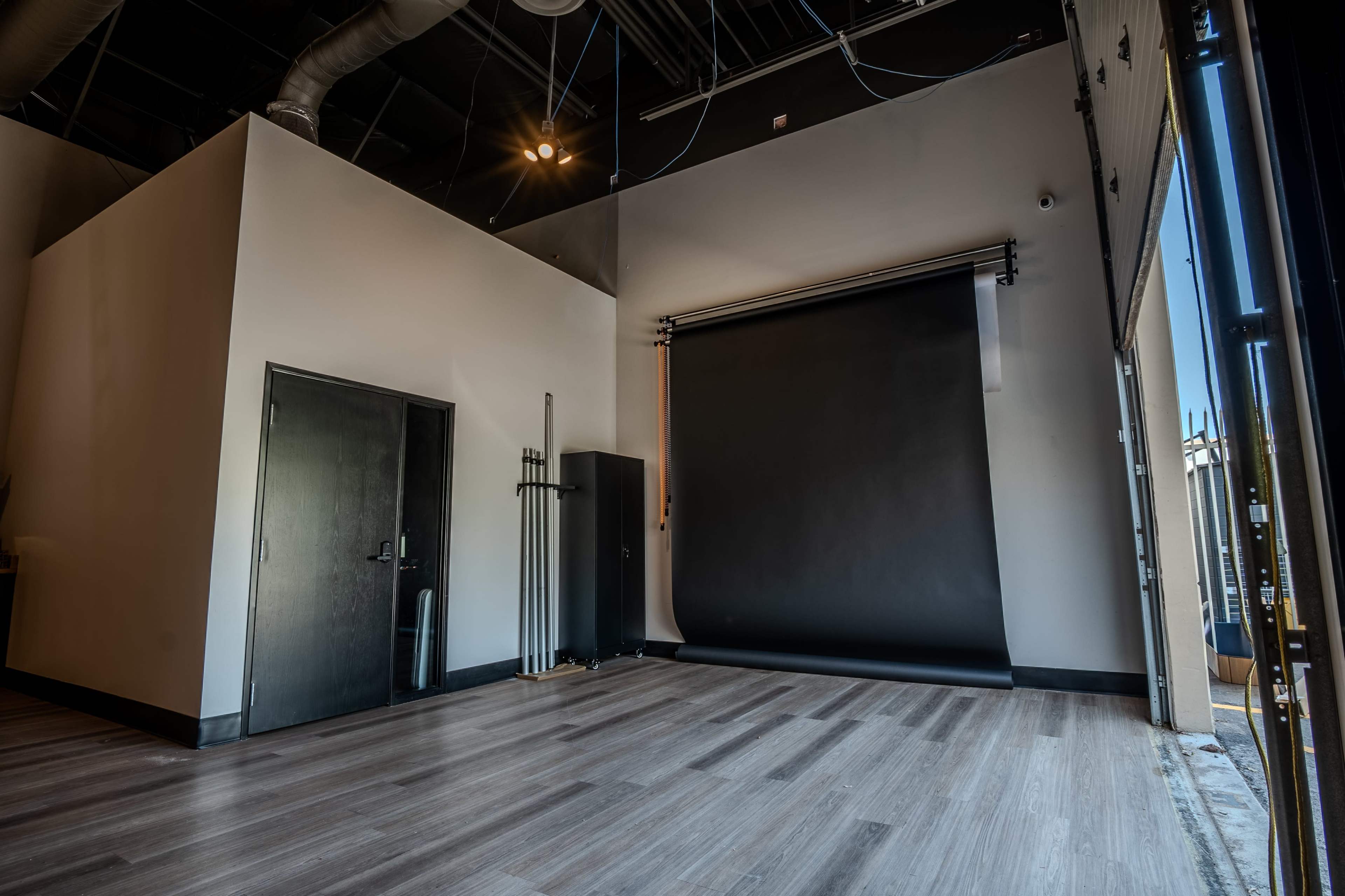 The image shows a spacious, empty photography studio featuring a black backdrop, hardwood flooring, and a door on one side.