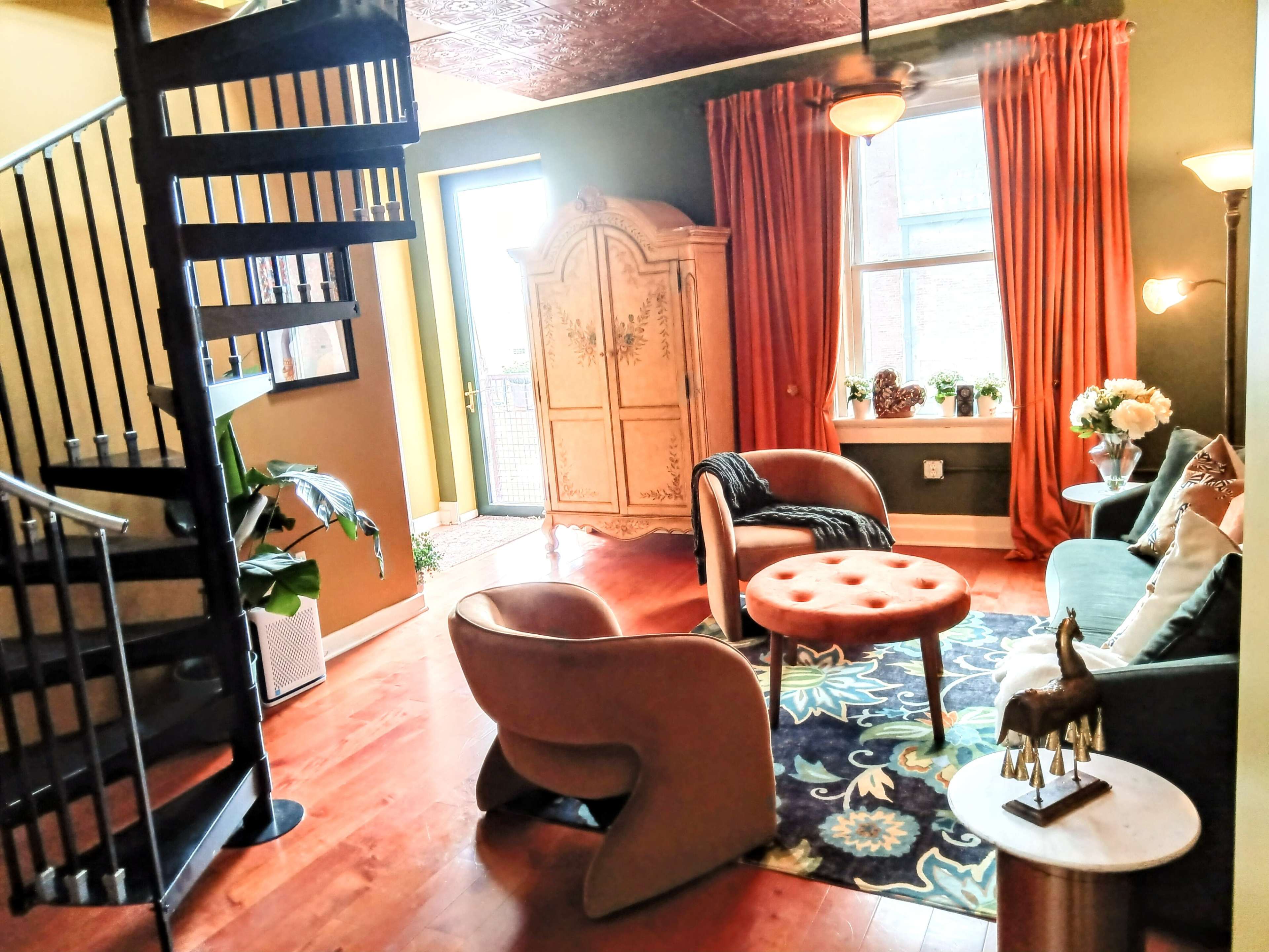A cozy living room with a spiral staircase, a circular coffee table, a pair of upholstered chairs, and large windows draped with orange curtains.