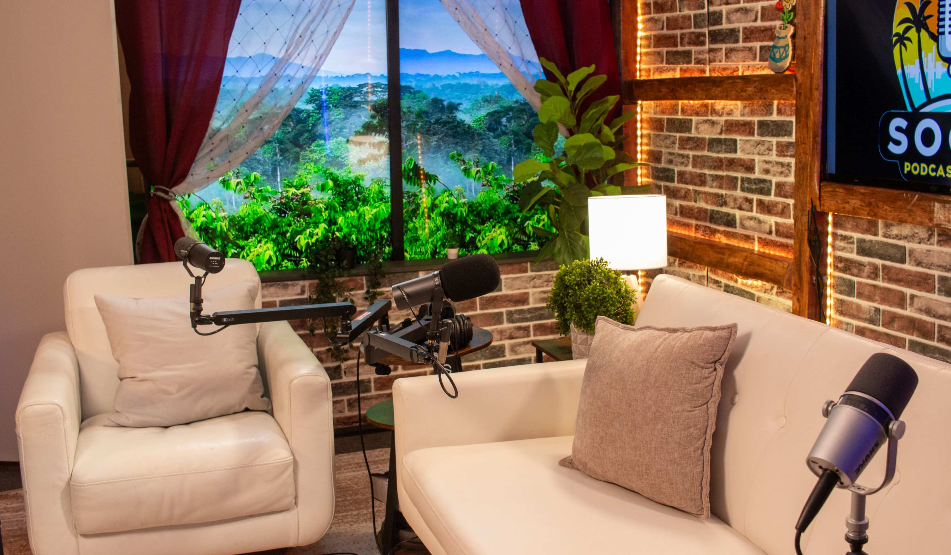 The image shows a cozy podcasting studio with two white chairs, microphones on stands, and a window displaying a green landscape.
