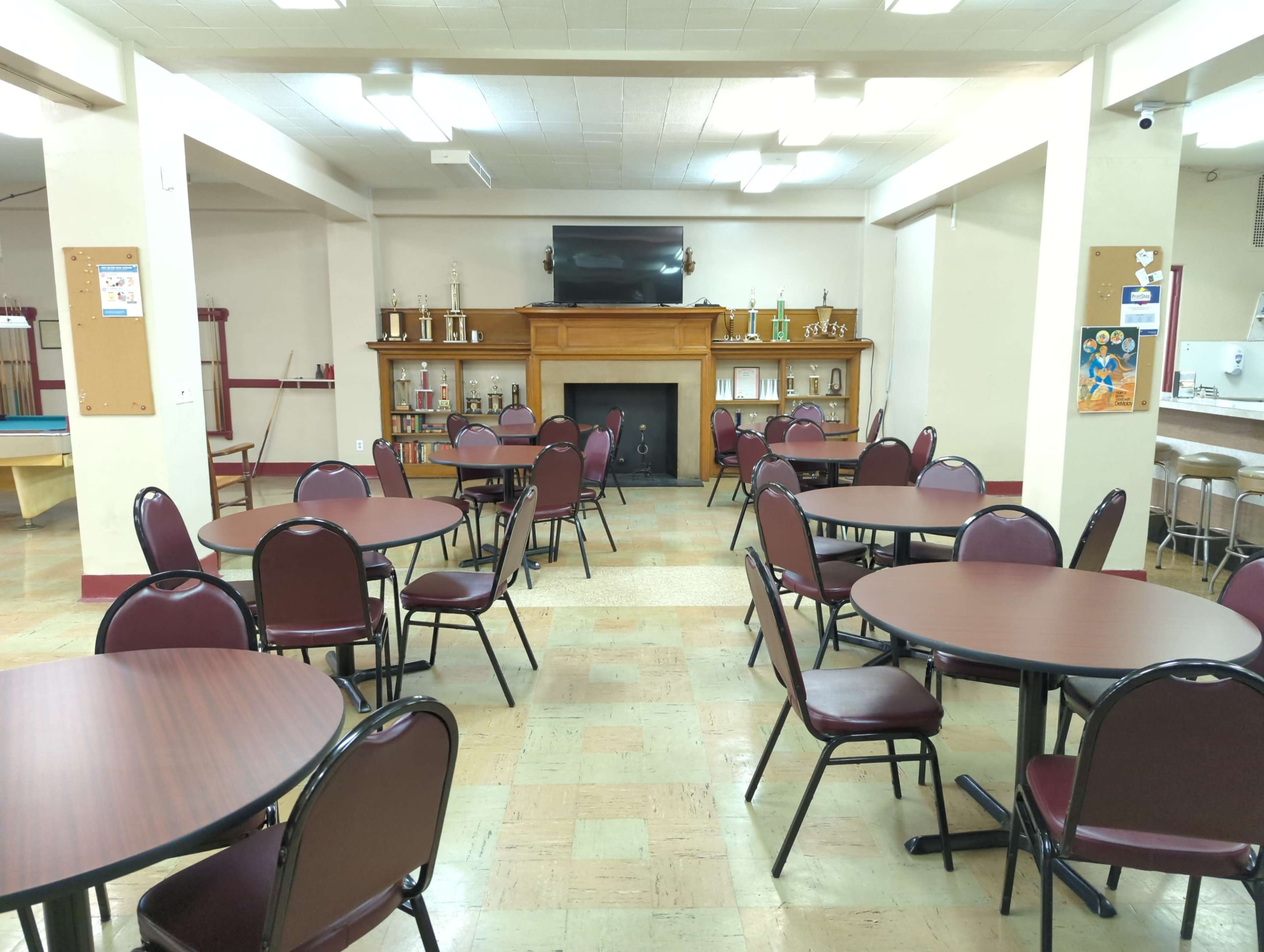 A spacious community room with several round tables, a fireplace, and shelves displaying trophies in the background.
