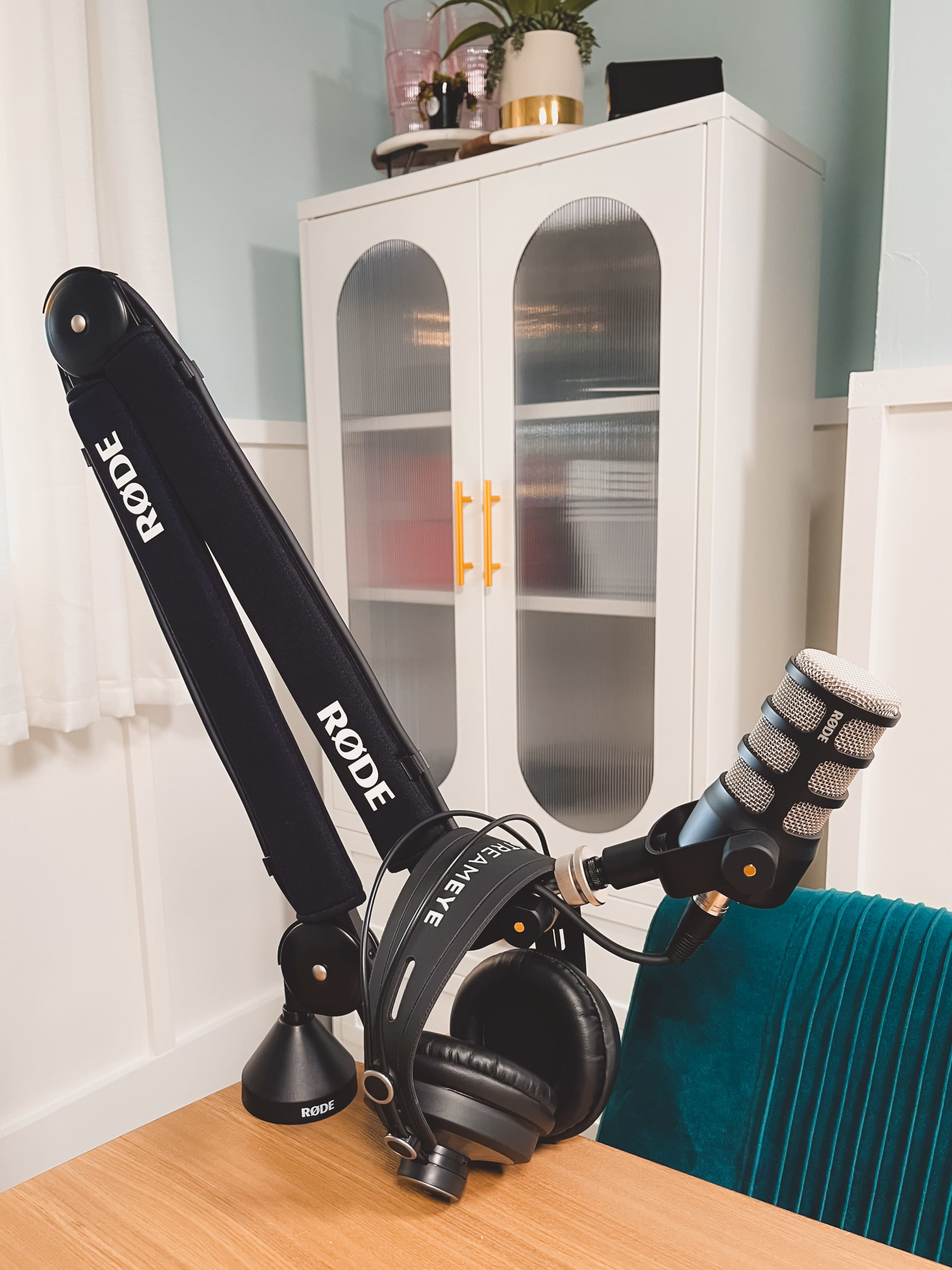 A microphone on a boom arm is positioned on a desk next to a pair of headphones, with a cabinet in the background.
