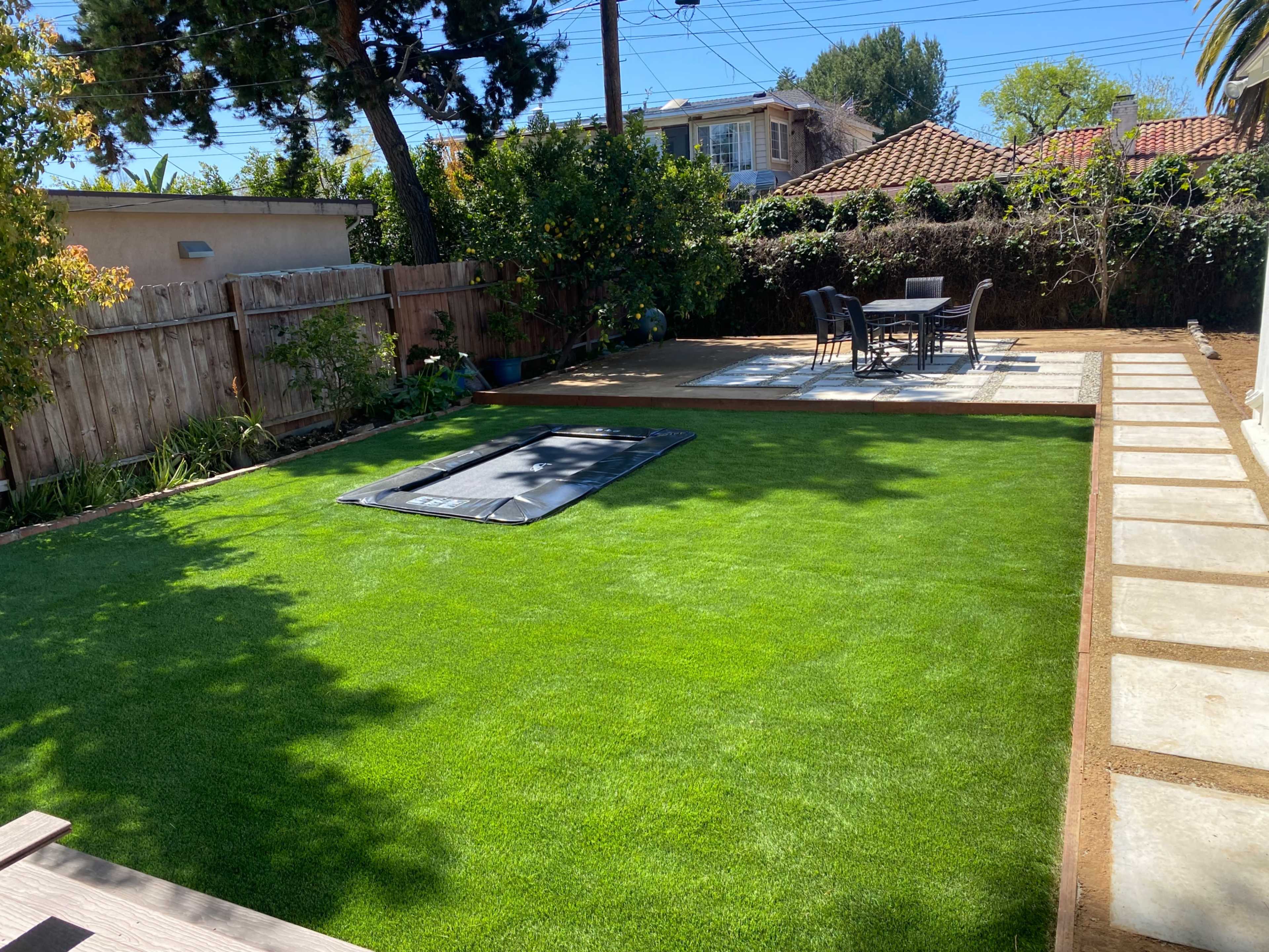 The image shows a backyard with a green lawn, a rectangular area covered by a tarp, and a patio set with a table and chairs.