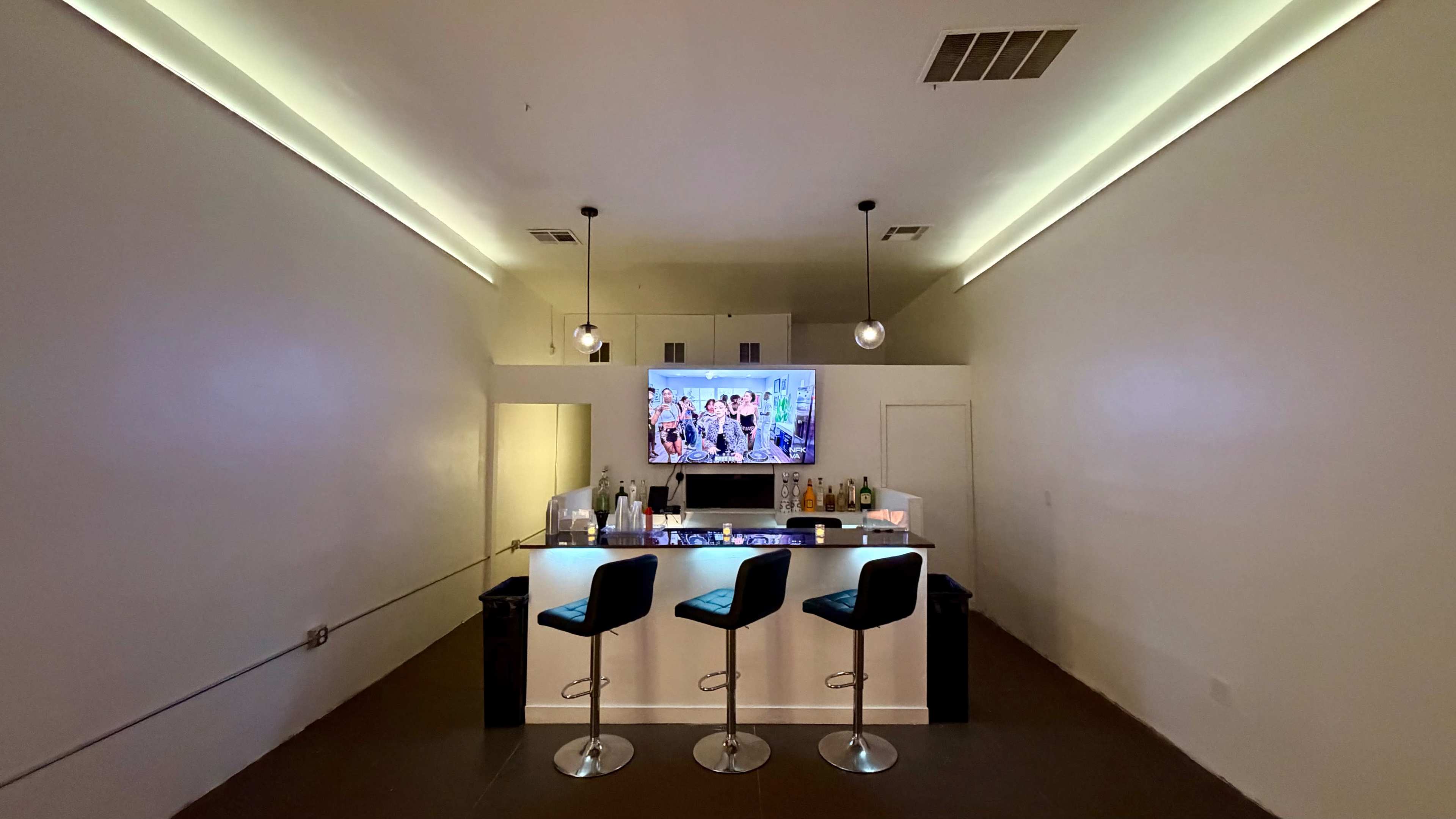 The image shows a modern bar area with a white counter, three high stools, a large television mounted on the wall, and various bottles displayed on the counter and shelves.