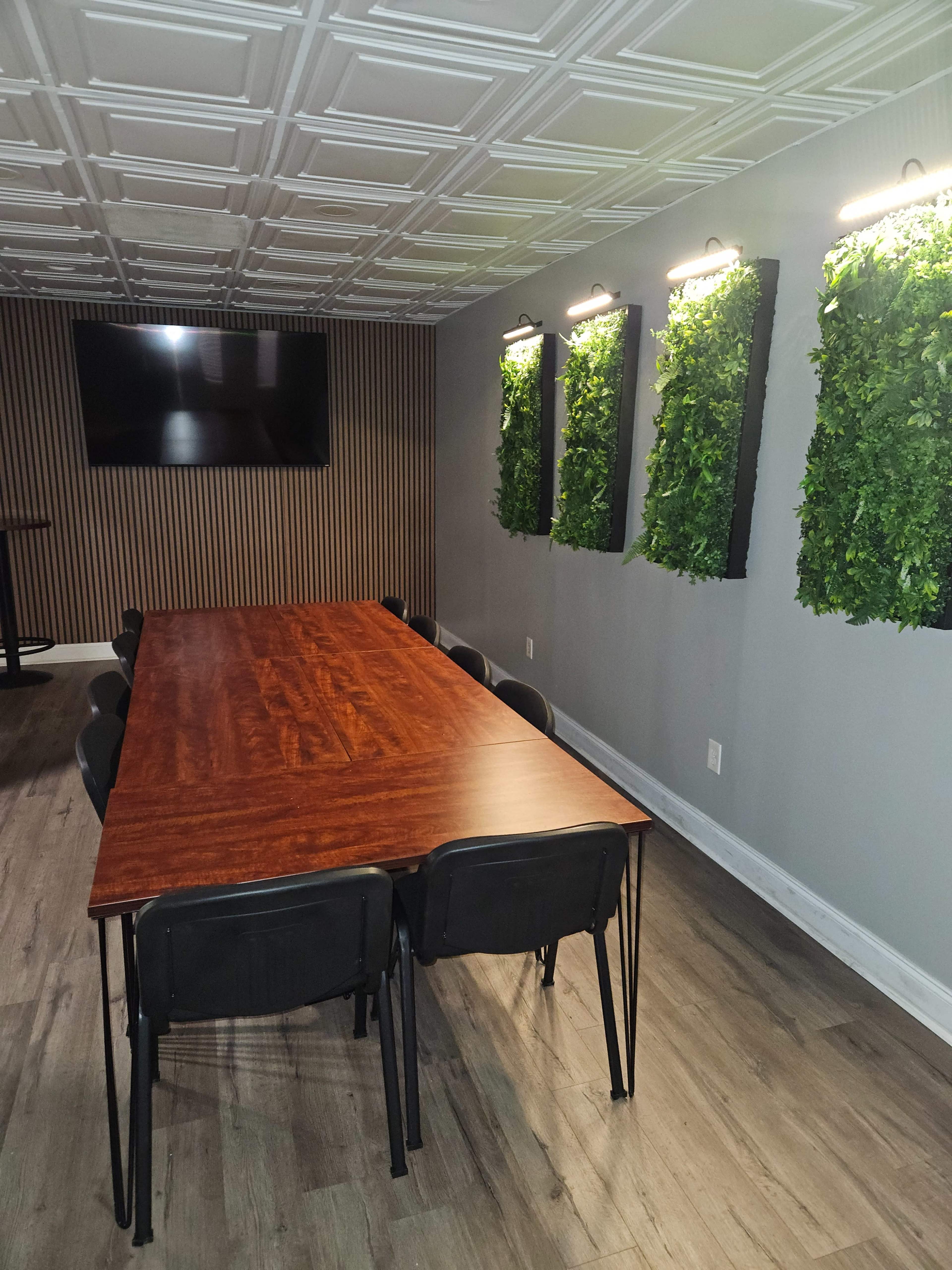The image shows a modern meeting room with a long wooden table surrounded by black chairs, green wall panels, and a wall-mounted TV.