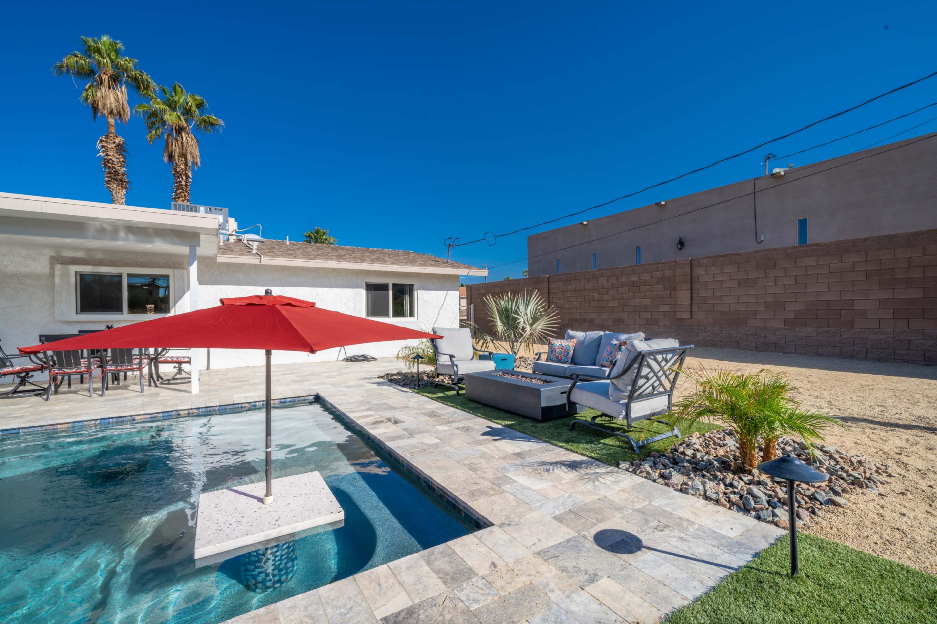 A swimming pool with a red umbrella, surrounded by a paved patio and decorative plants, is situated next to a house.