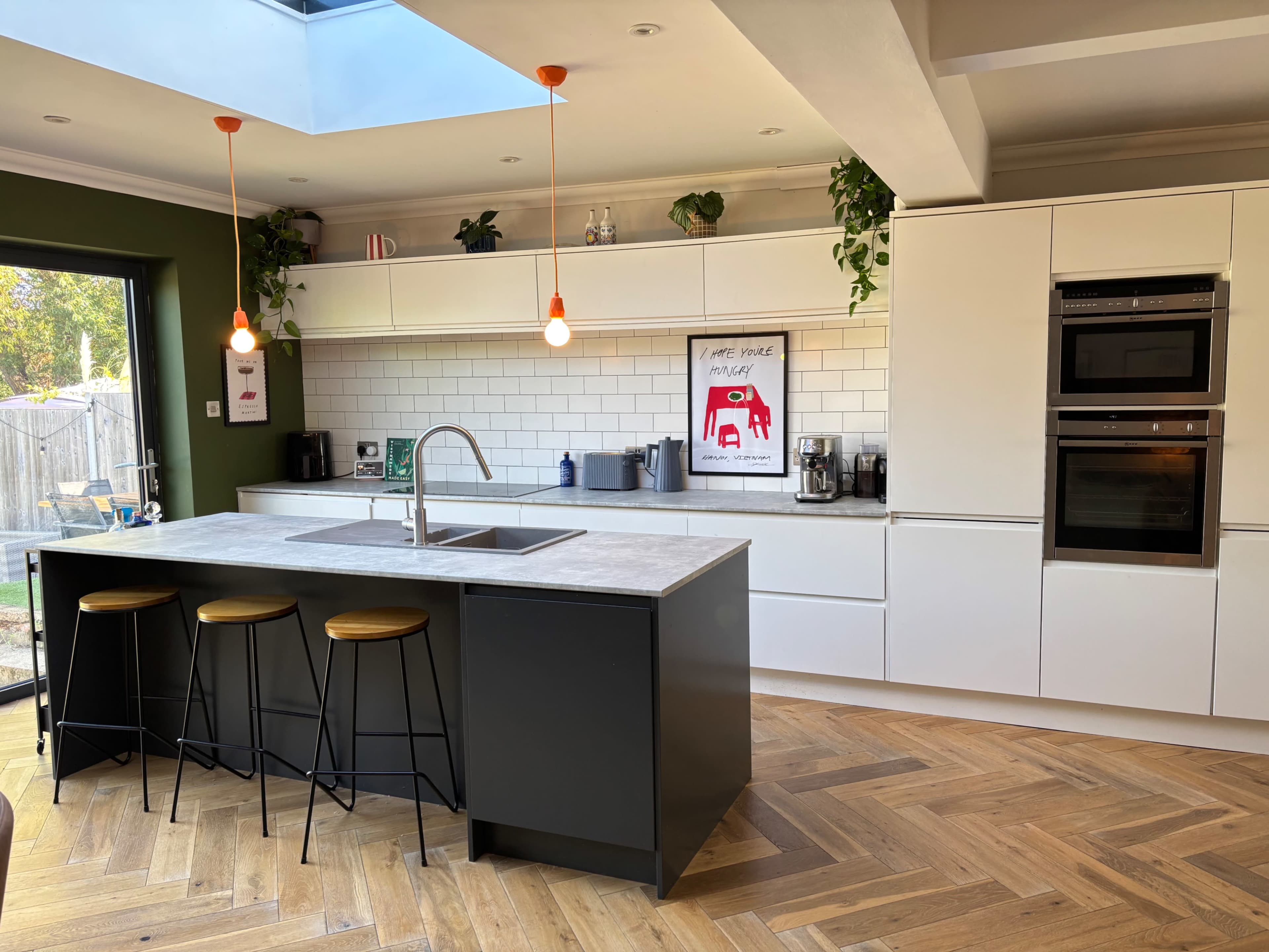 The image features a modern kitchen with a central island, three stools, and a skylight, accented by green walls and various potted plants.