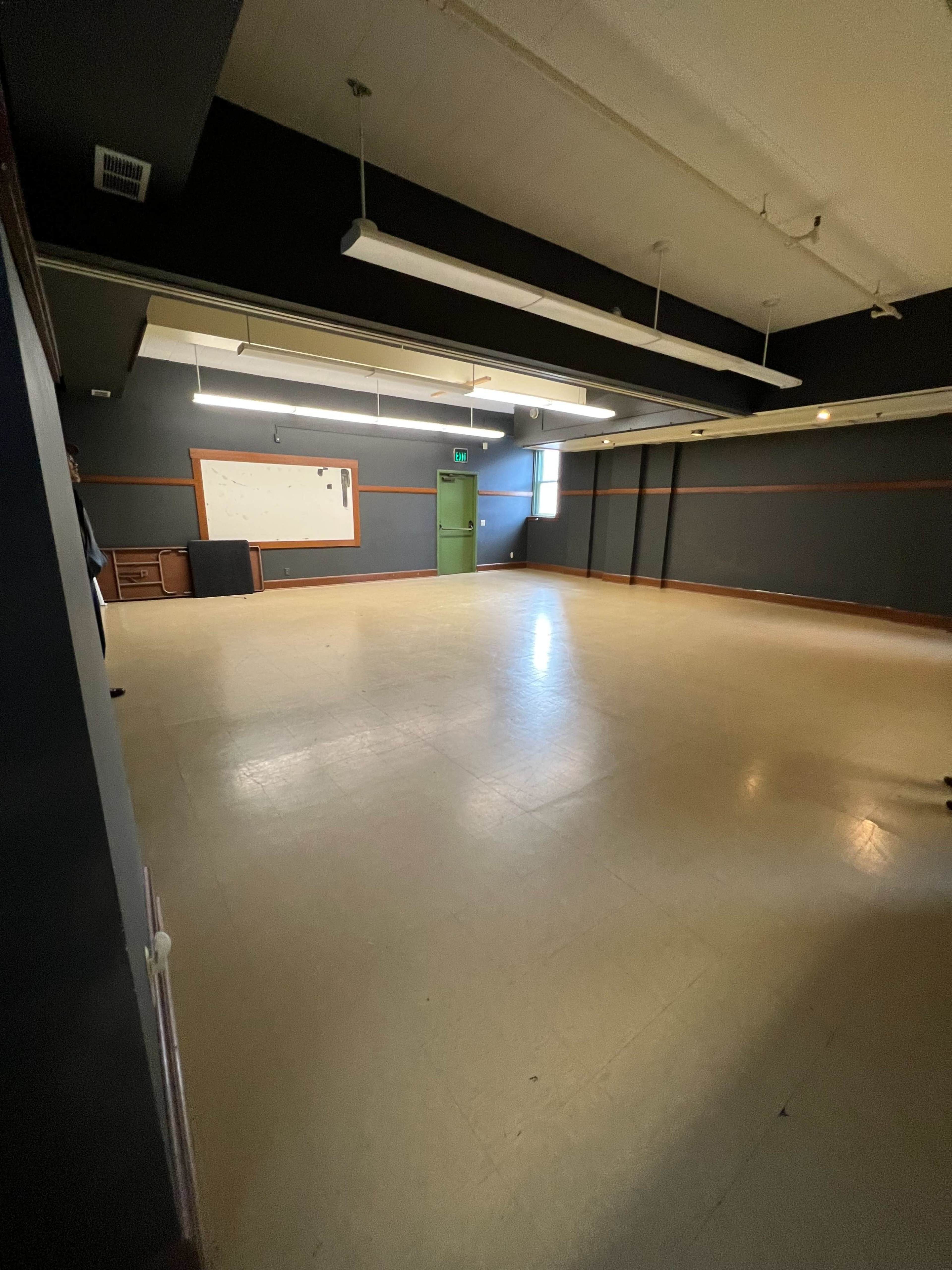 An empty room with light-colored flooring, dark walls, and a green door, featuring overhead lighting and a whiteboard on one wall.