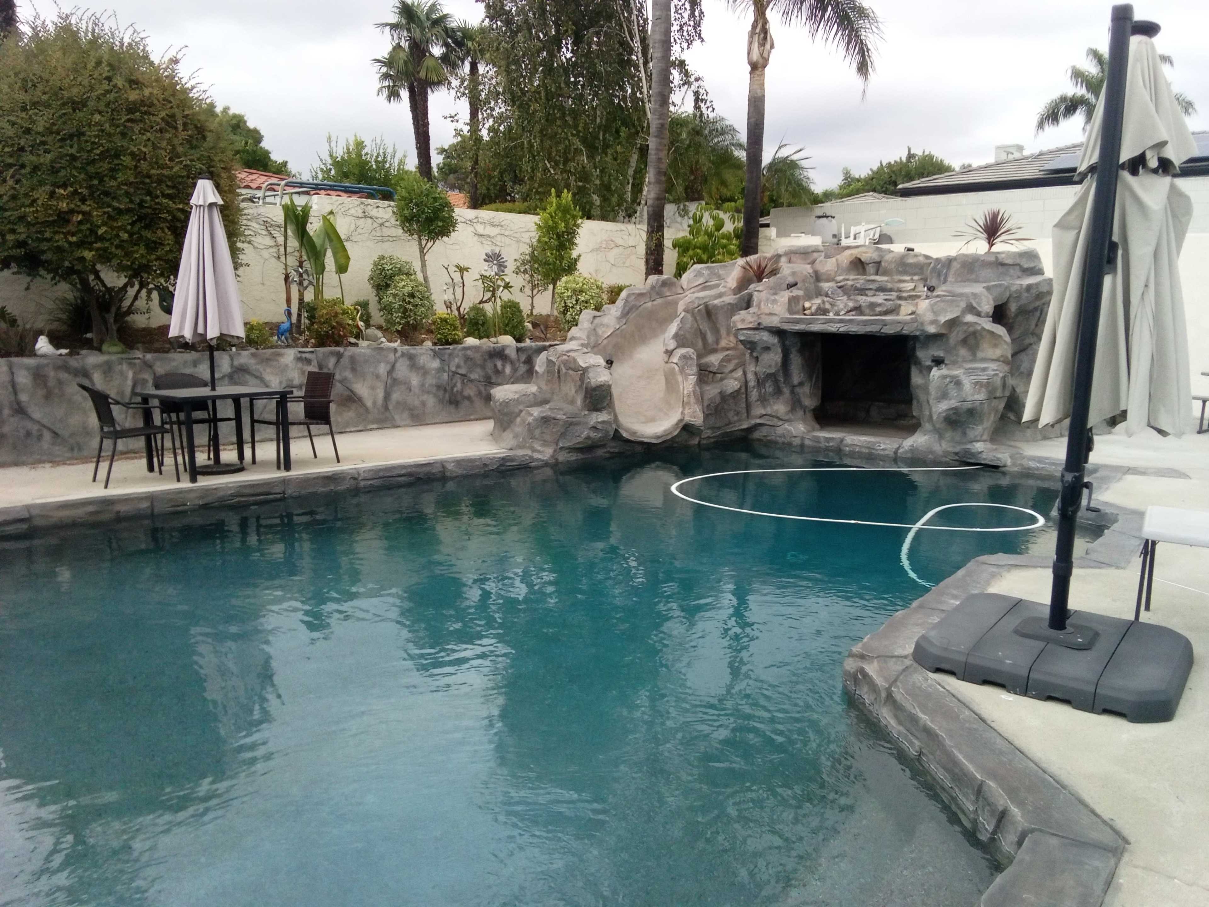 The image shows a swimming pool surrounded by rock formations, featuring a waterslide and a lounge area with patio furniture under a sunshade.