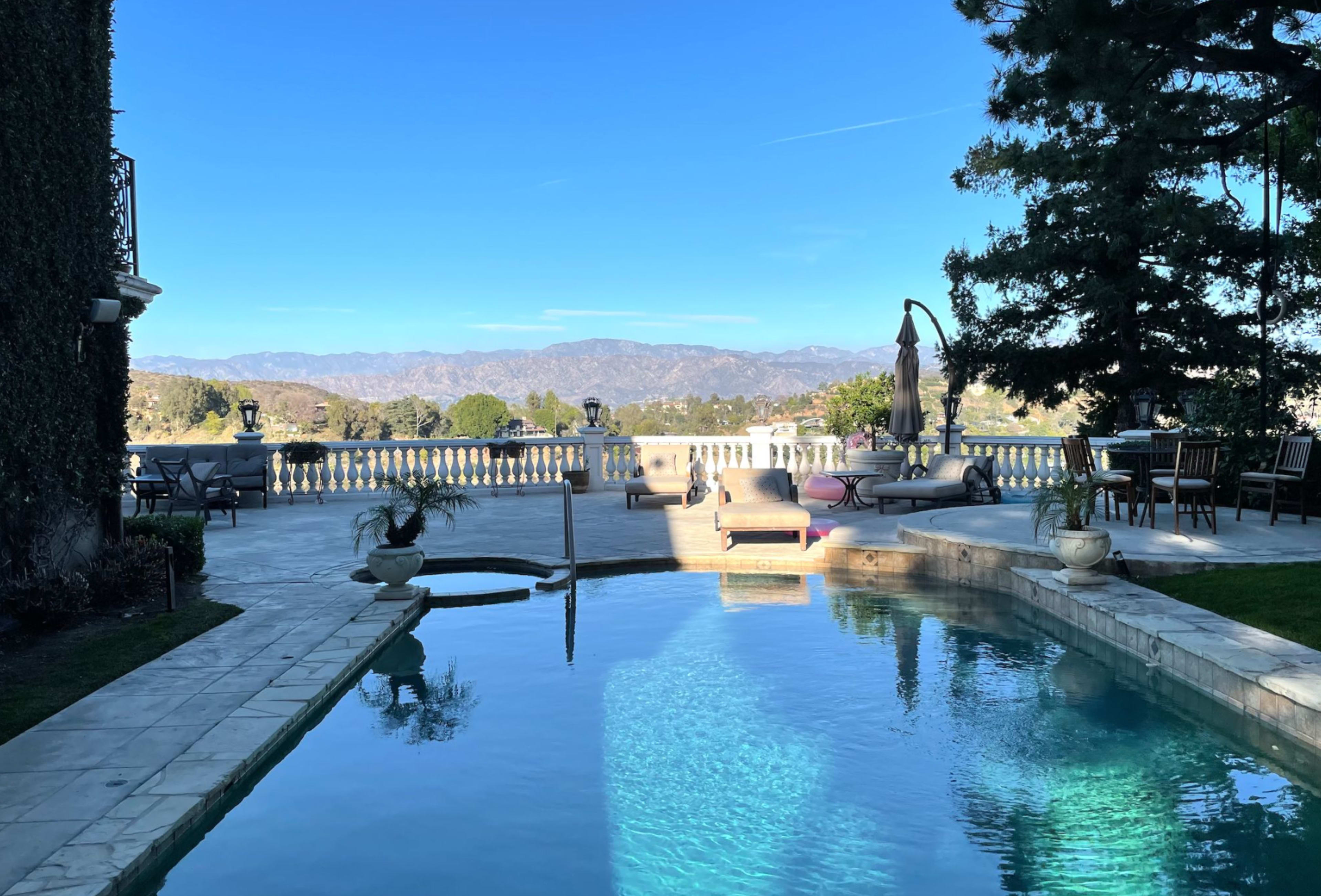 The image shows a swimming pool surrounded by a patio with furniture and a view of distant mountains under a clear blue sky.