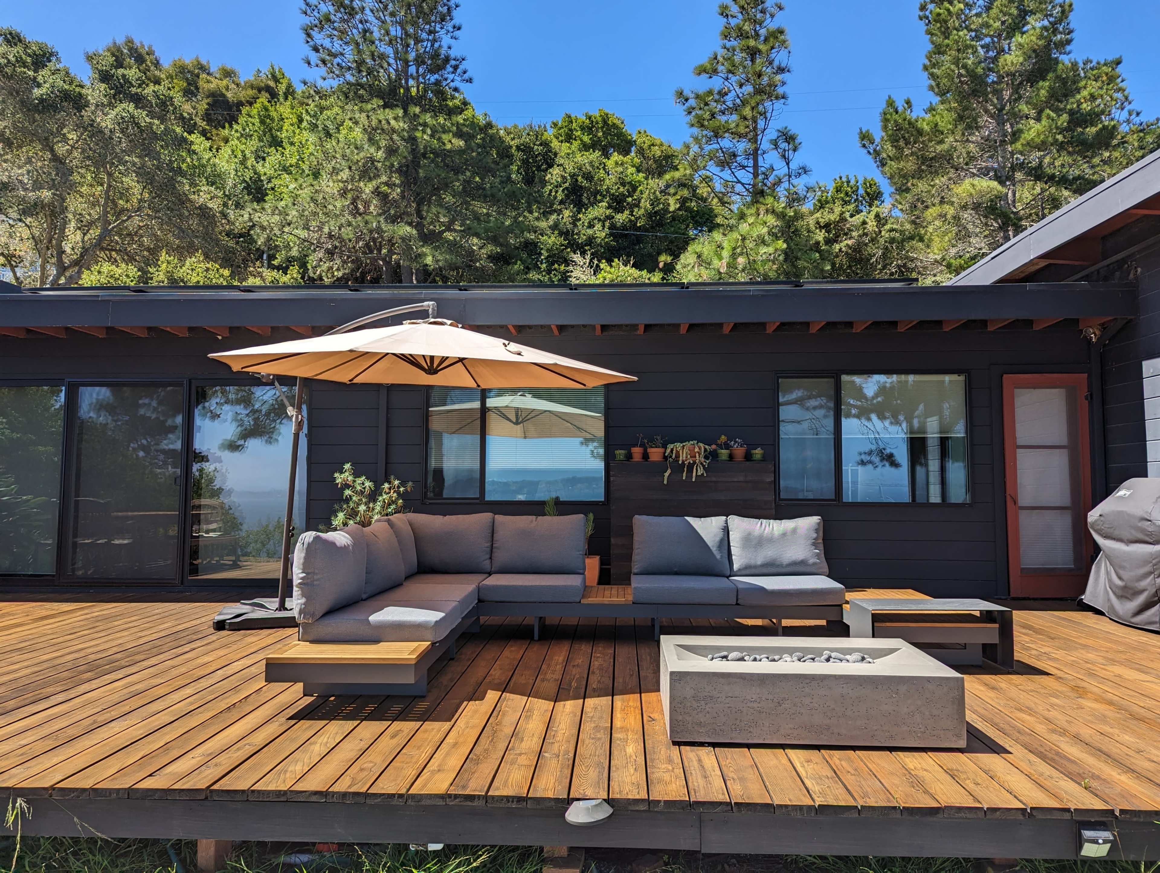 A wooden deck features a sectional sofa, a large umbrella, and a fire pit in front of a house surrounded by trees and a view of the water.