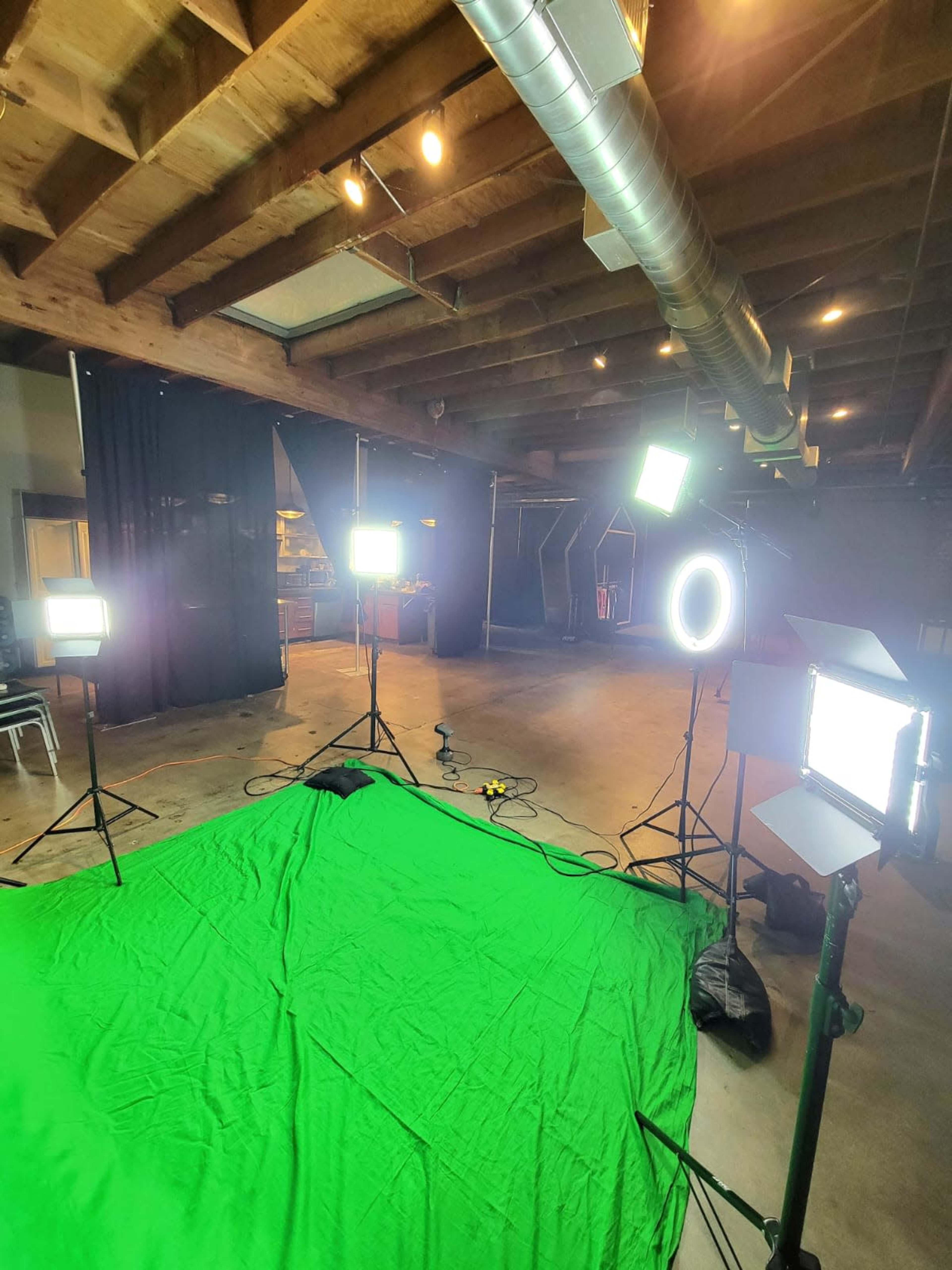 The image shows a green screen setup in a spacious indoor area, surrounded by multiple bright lights on stands, with black curtains in the background.