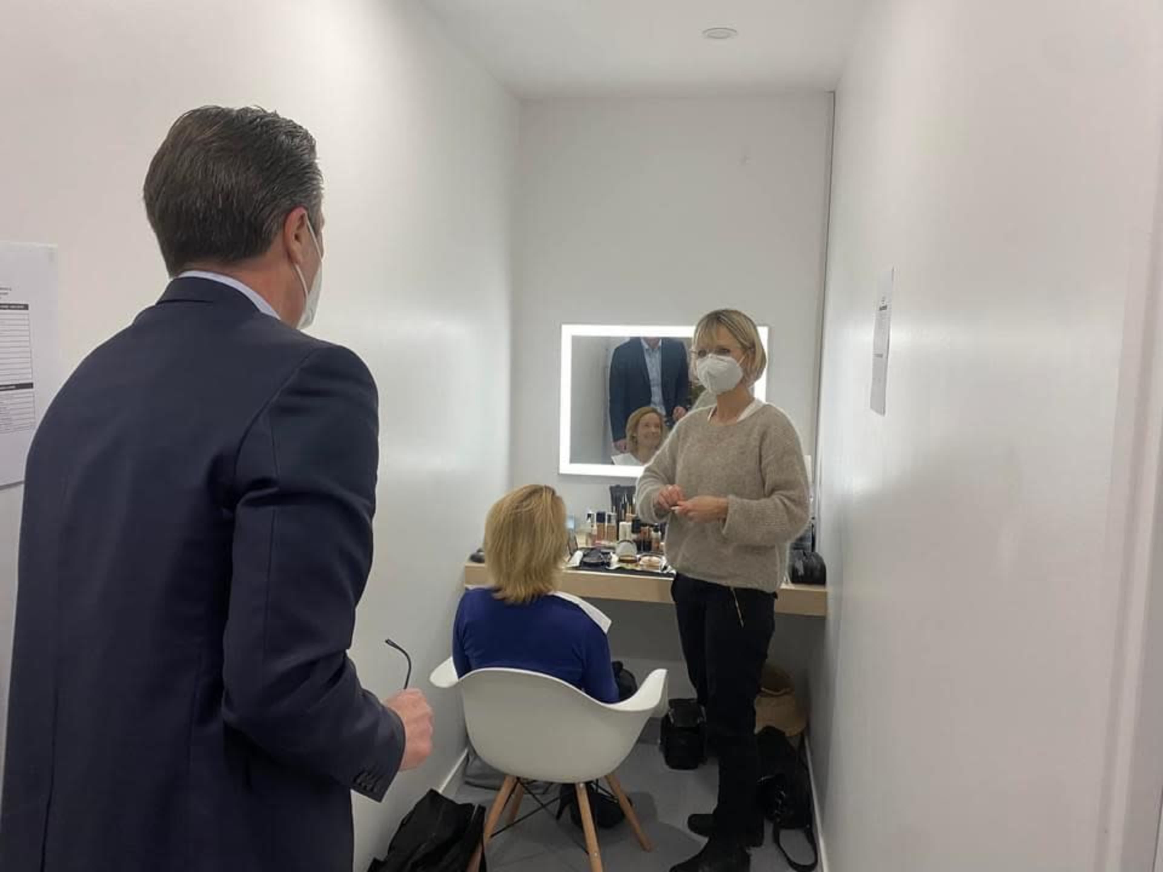 A man in a suit stands talking to a woman in a beige sweater while another woman sits in a chair in front of a makeup station in a small, well-lit room.